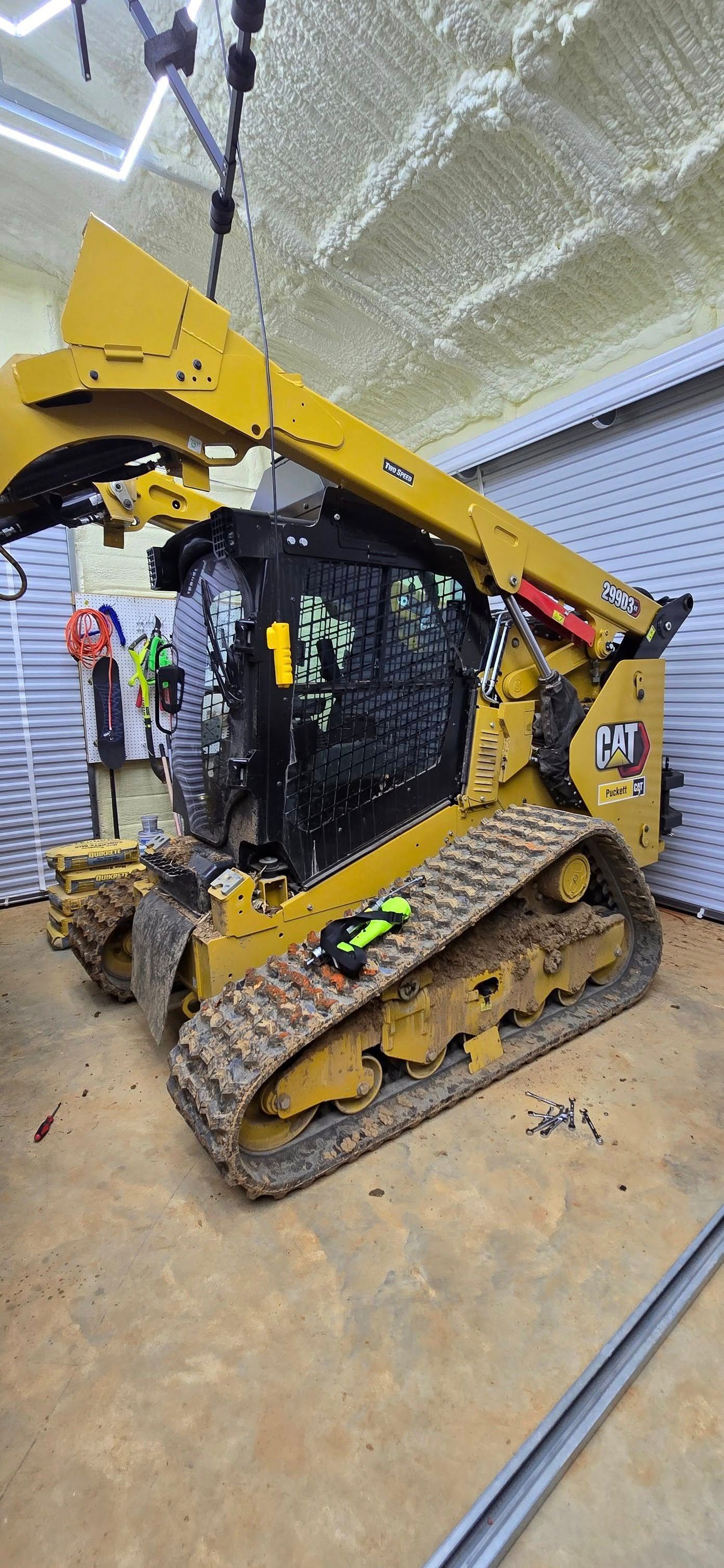 Yellow track skid steer in a workshop. The boom is raised, showing black engine components.