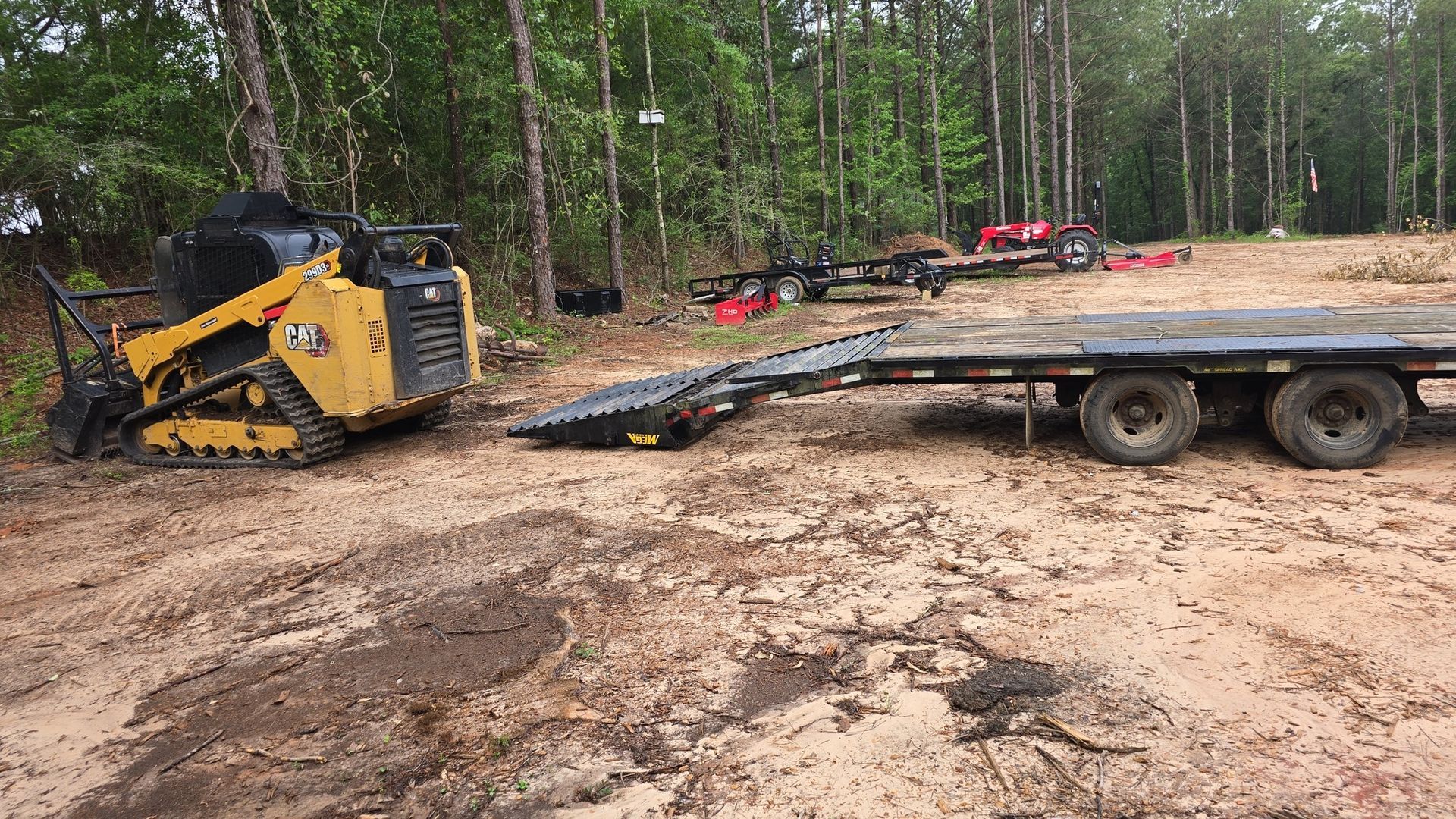 A yellow tracked skid steer loading onto a flatbed trailer in a muddy clearing, forest in background.