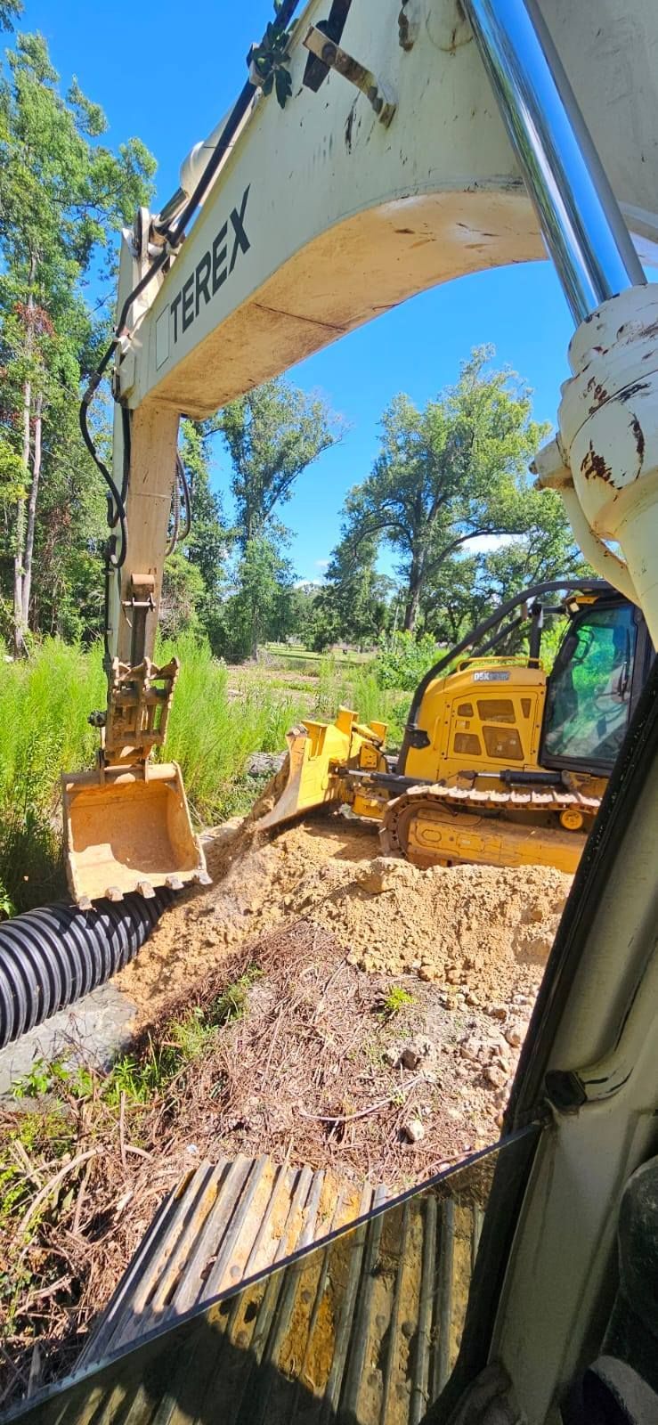An excavator and a bulldozer work on a construction site next to a culvert. Trees and blue sky in the background.