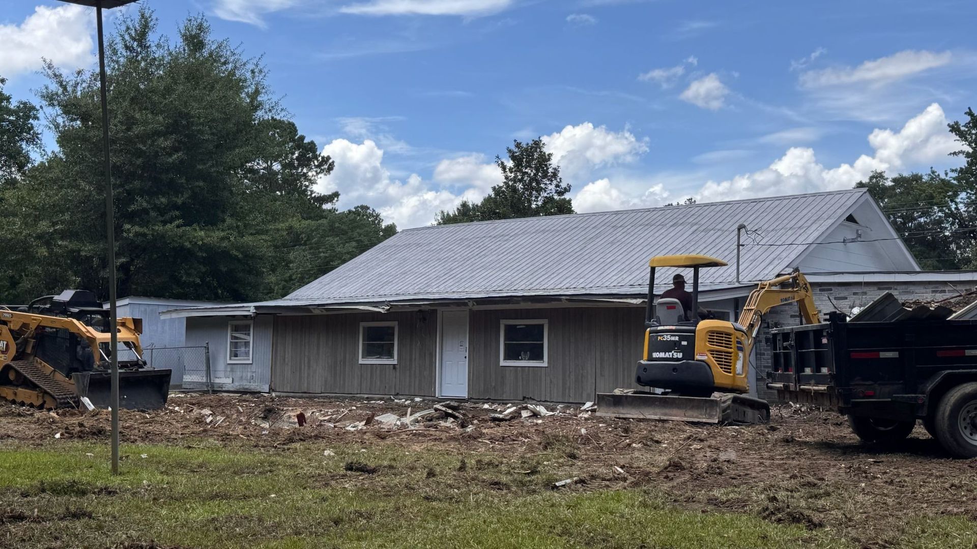 Construction site: Excavator and bulldozer near a building with a metal roof. Cloudy sky.
