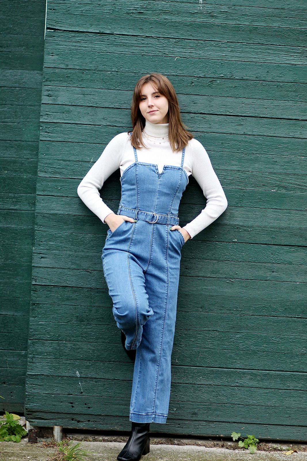 A person wearing a white turtleneck and blue denim jumpsuit leaning against a dark green wooden wall.