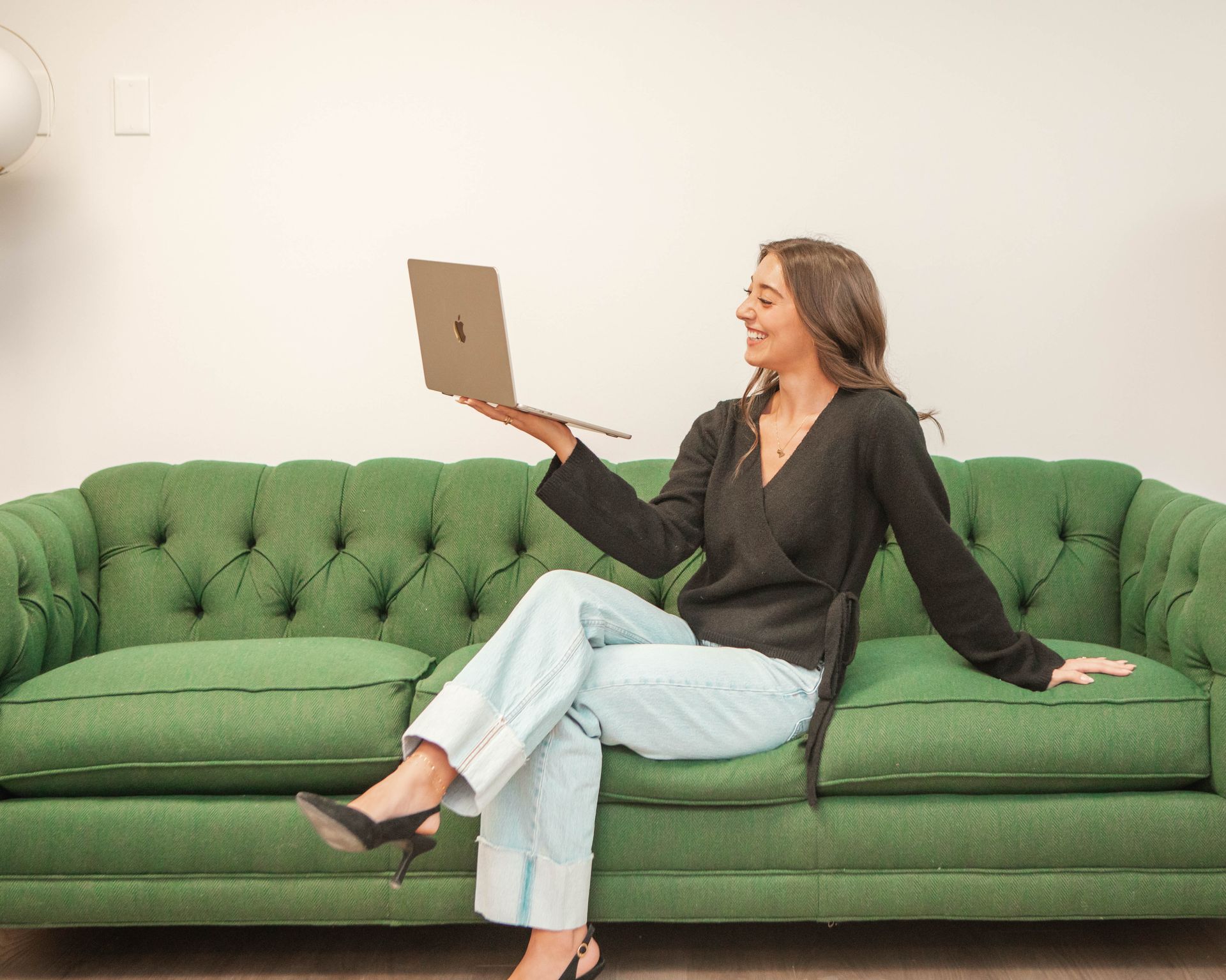 A person sits on a green tufted sofa, smiling and holding an open laptop in one hand.
