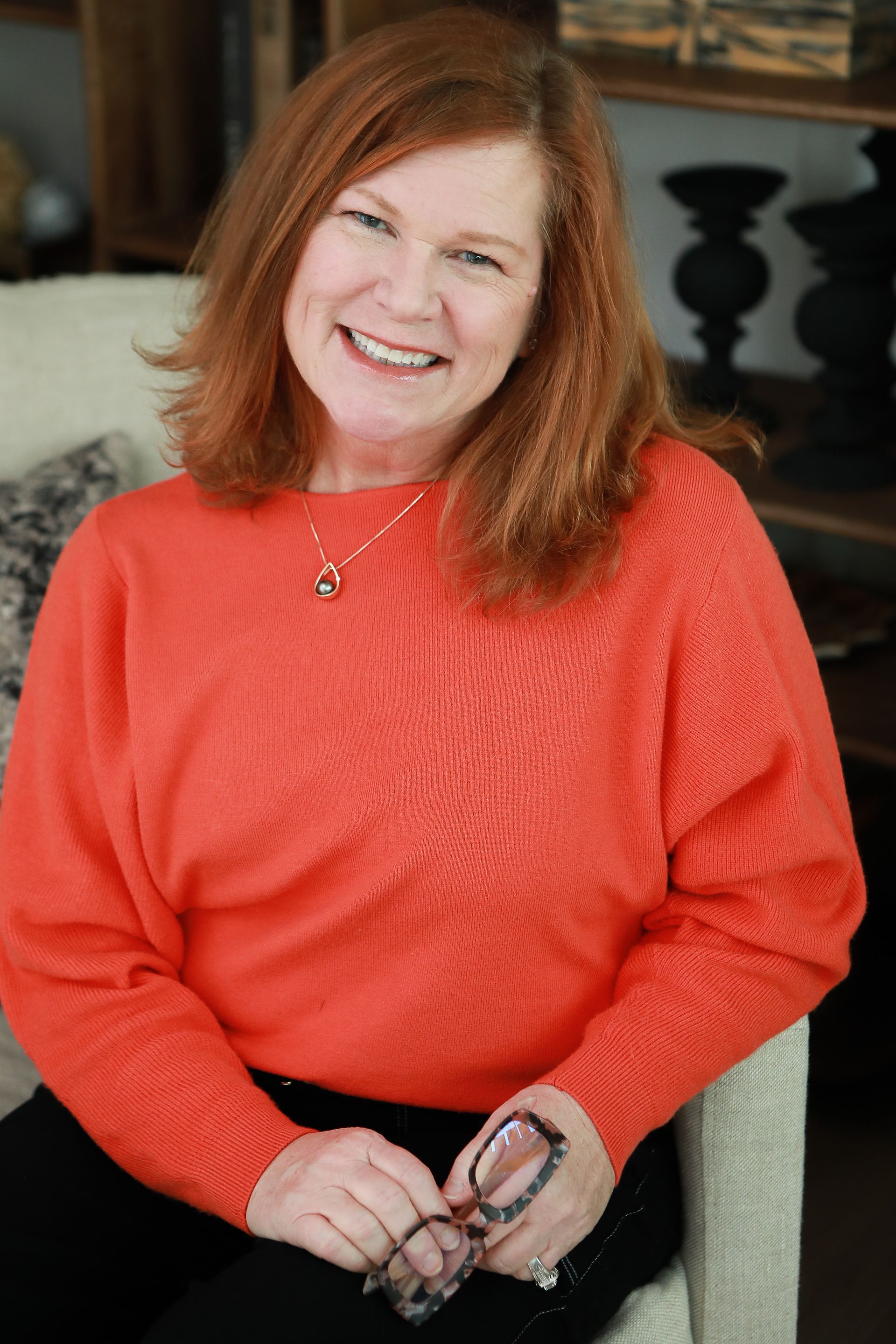 A person with auburn hair smiles, wearing an orange sweater and a necklace, holding glasses in their lap.
