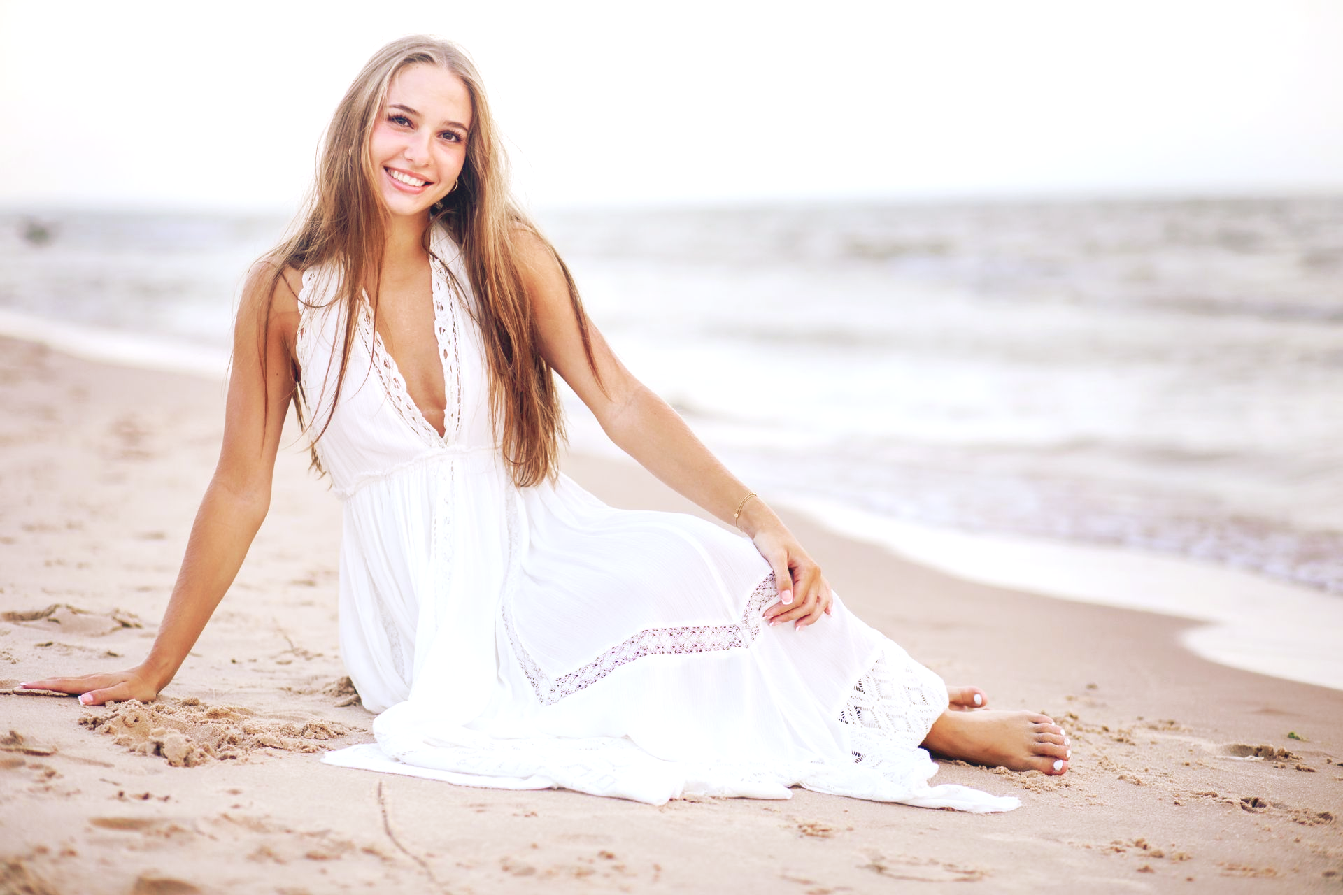 A smiling person in a long white dress sits on a sandy beach by the ocean.