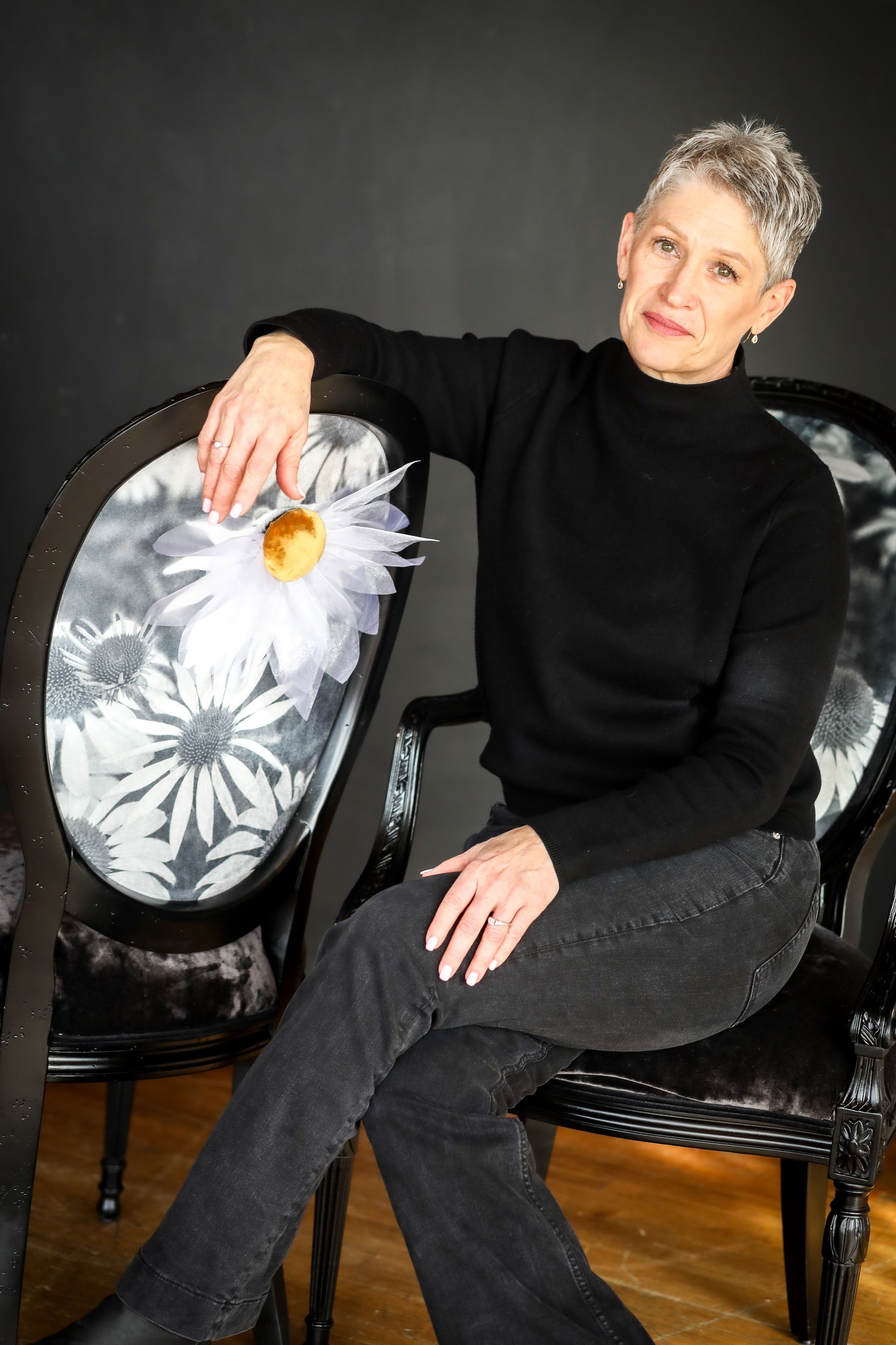 A person with short hair, wearing a black sweater and jeans, sits on a chair with a daisy-patterned backrest.