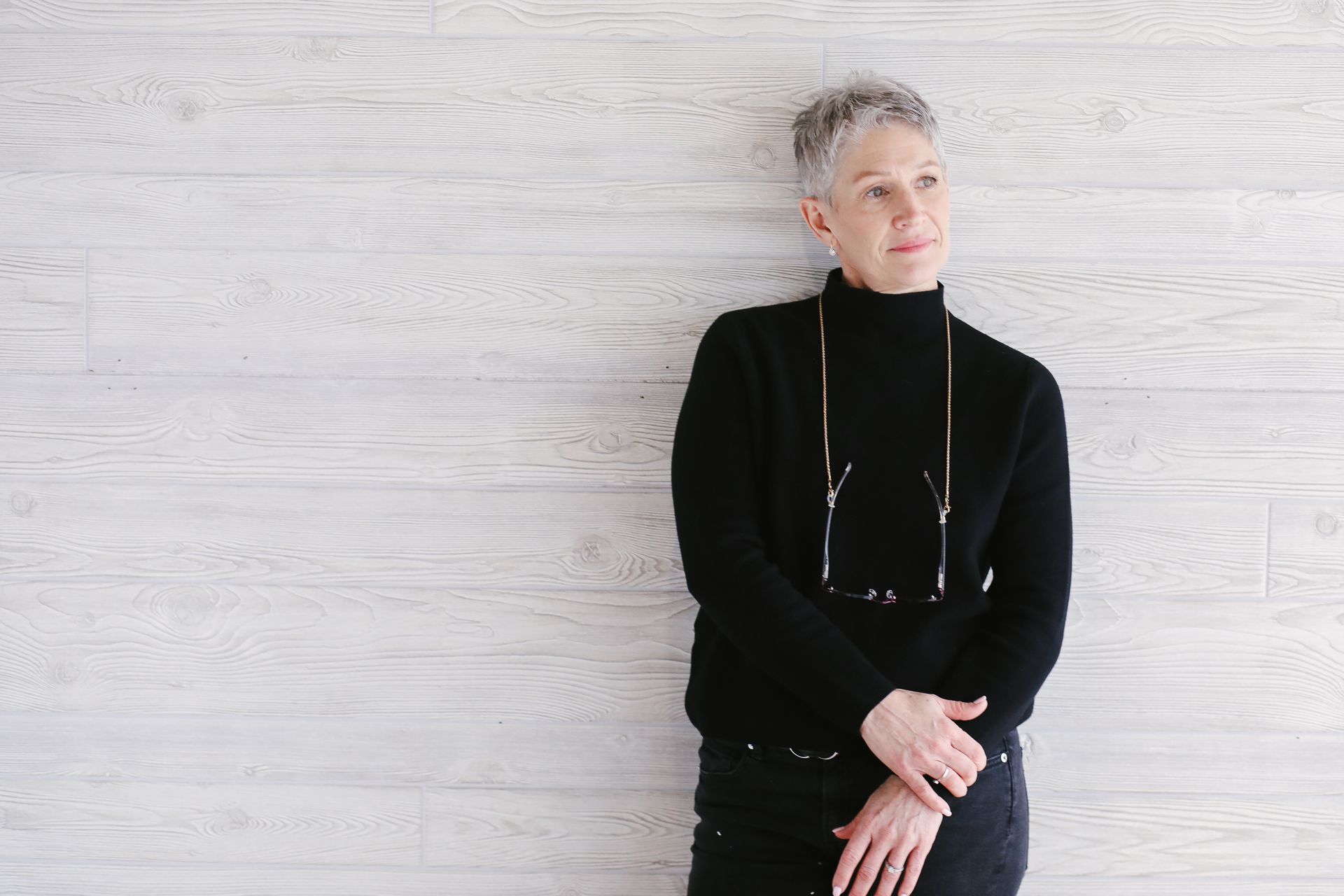 A person with short grey hair wearing a black turtleneck and glasses around their neck, leaning against a white wall.
