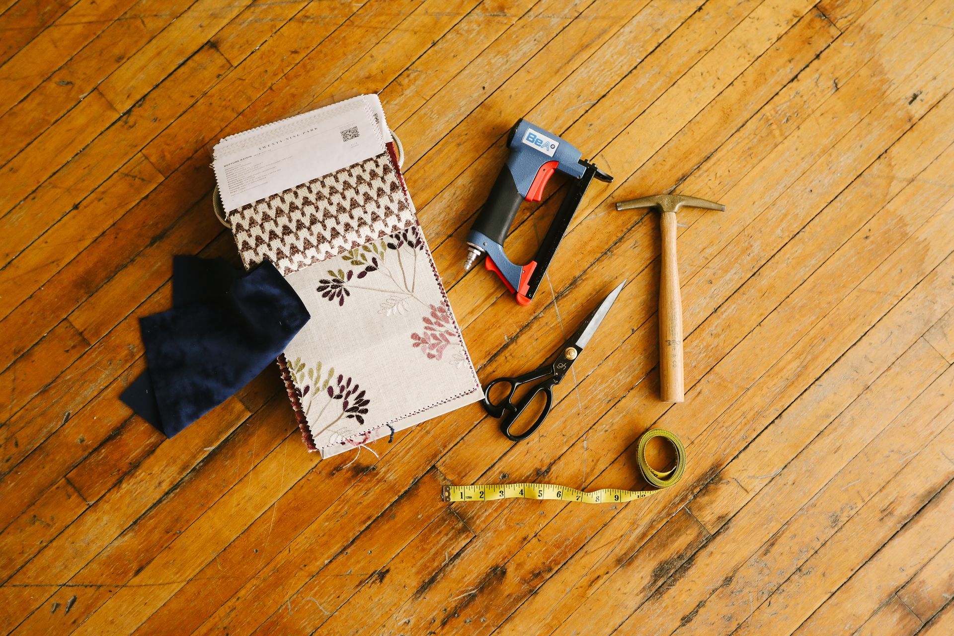 Upholstery tools, including a staple gun, hammer, scissors, fabric swatches, and a tape measure on a wooden floor.