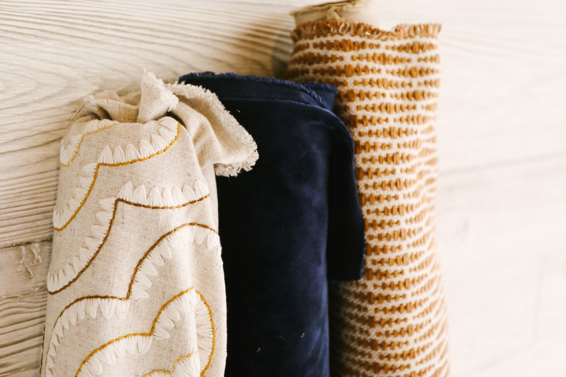 Three fabric rolls standing against a light-colored, wood-textured wall: one cream with gold embroidery, one navy, one tan.