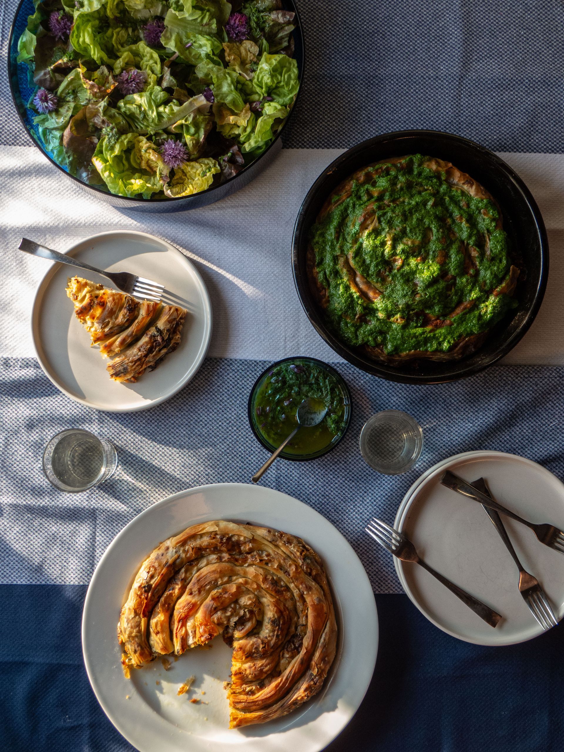 Three plated savory dishes and salads arranged on a blue-checkered tablecloth with small bowls of sauce, don't call me burek
