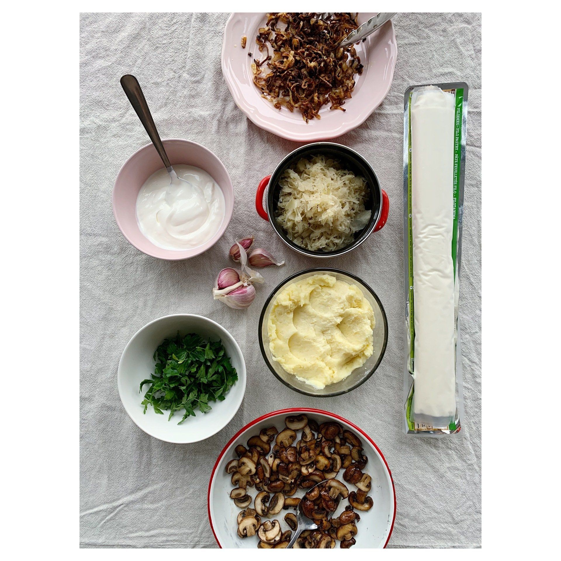 Assorted baking ingredients in bowls: phyllo dough, sour cream, sauerkraut mushrooms, garlic, herbs on a gray cloth.