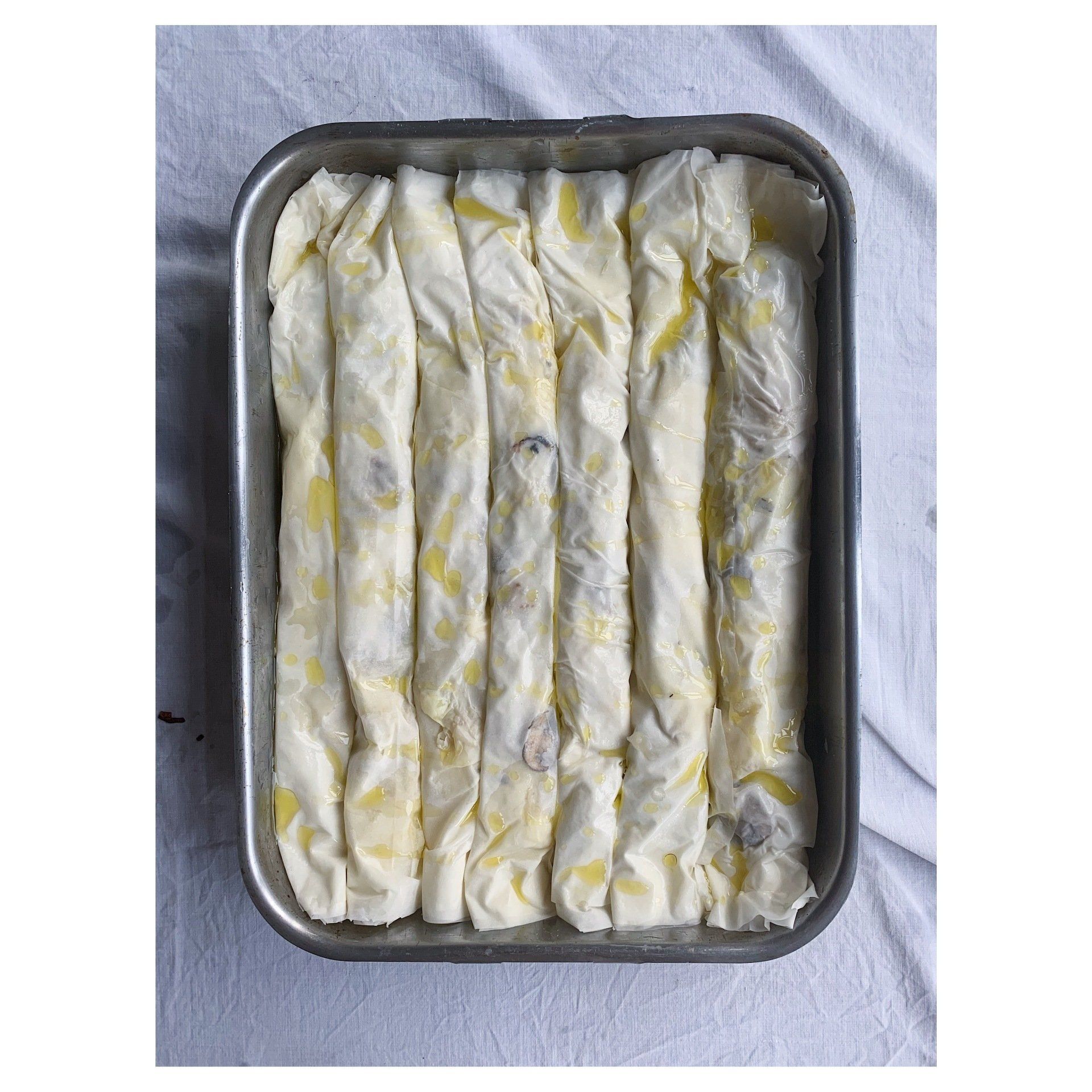 Tray of rolled pastry dough brushed with oil, arranged in rows on a white cloth background