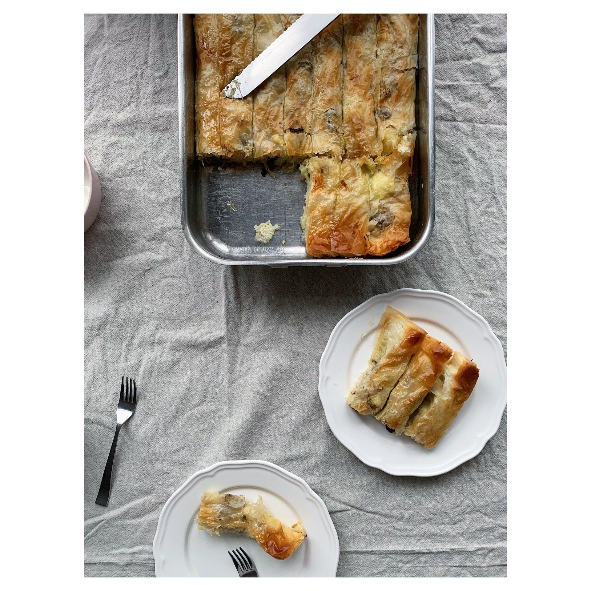 Baked savoury pastry in a tray with two sliced pieces on white plates on a gray tablecloth.