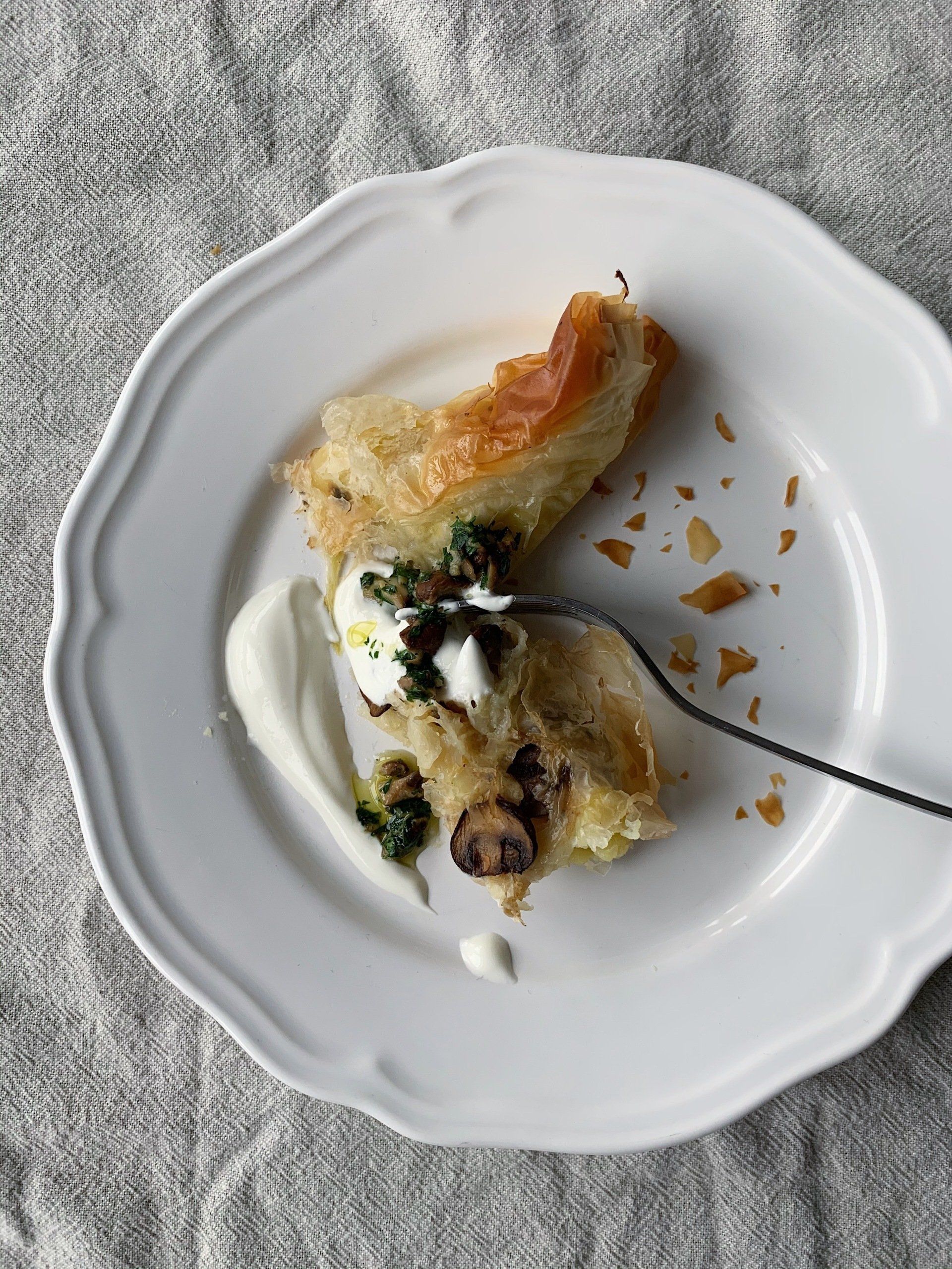 Partially eaten burek on a white plate with sour cream and crumbs on a gray textured tablecloth