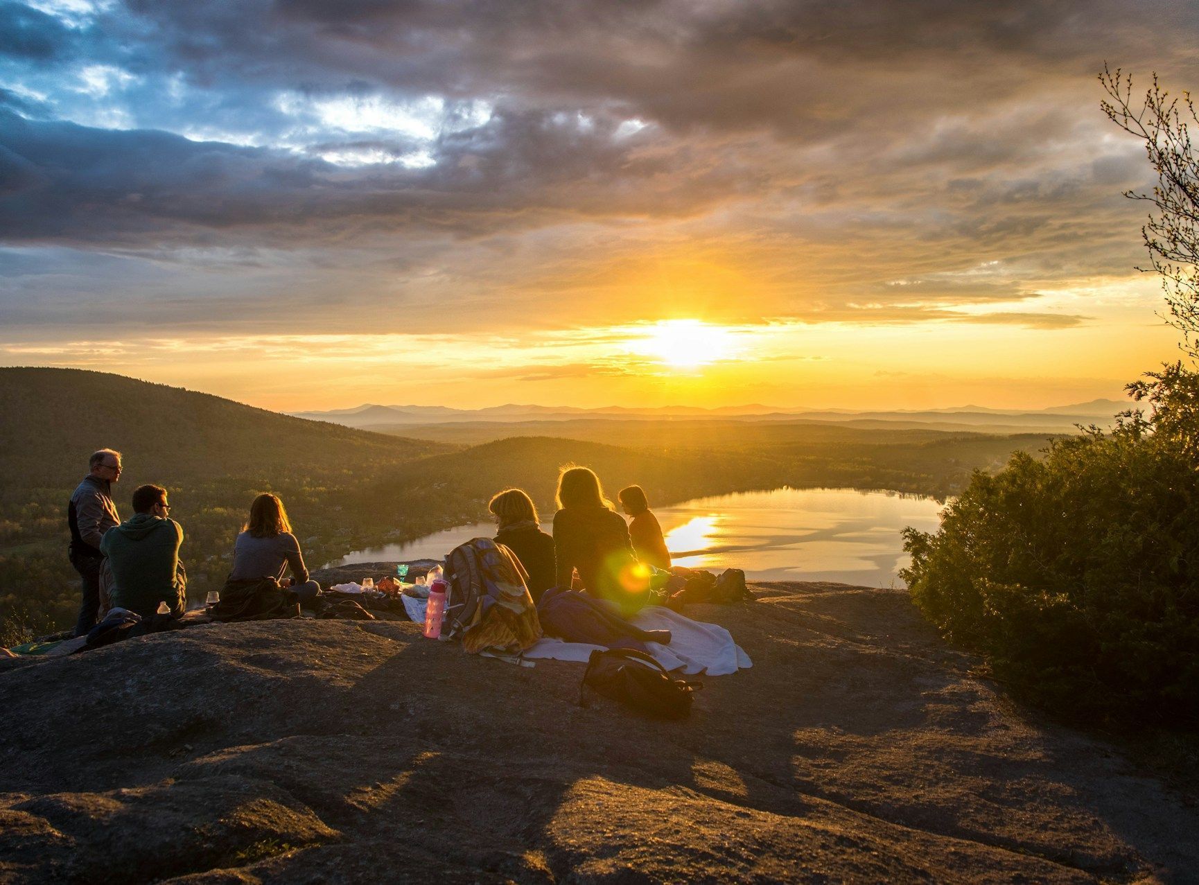 A group of people are sitting on top of a mountain watching the sunset over a lake.