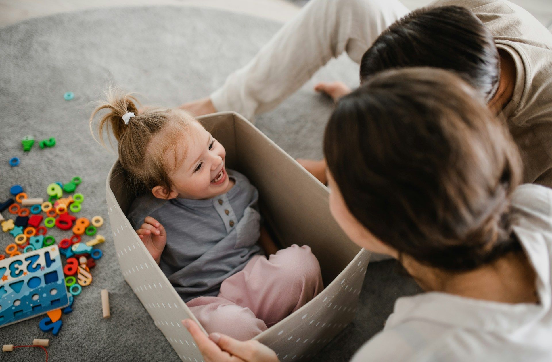 A little girl is sitting in a cardboard box on the floor.
