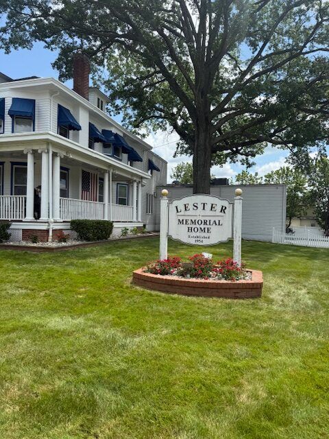Lester Memorial sign in front of a white house with blue awnings and large trees on a grassy lawn
