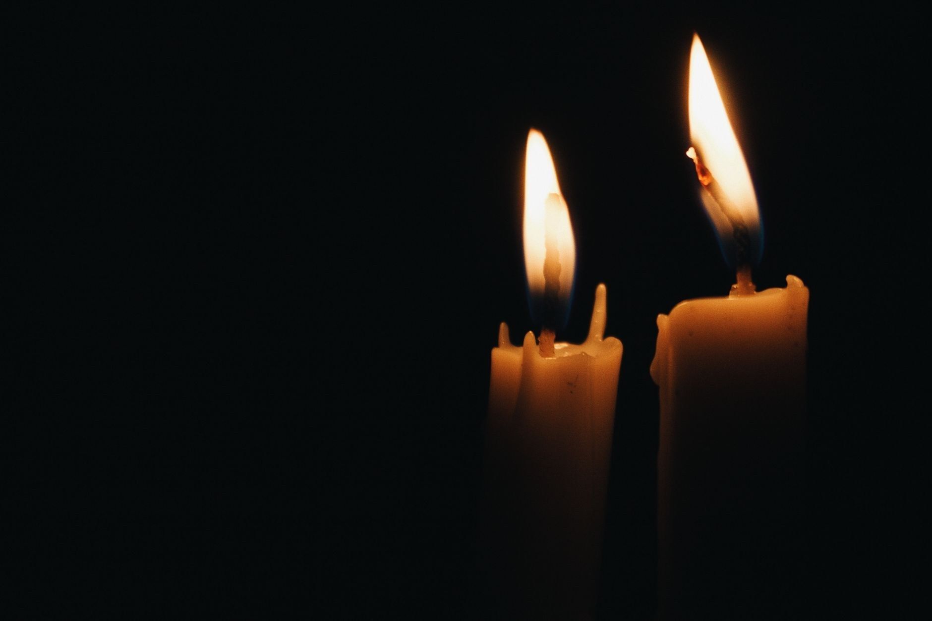 Two lit candles glow against a completely black background, their flames the only source of light in the frame.