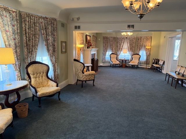 Spacious carpeted living room with floral curtains, upholstered chairs, and a chandelier