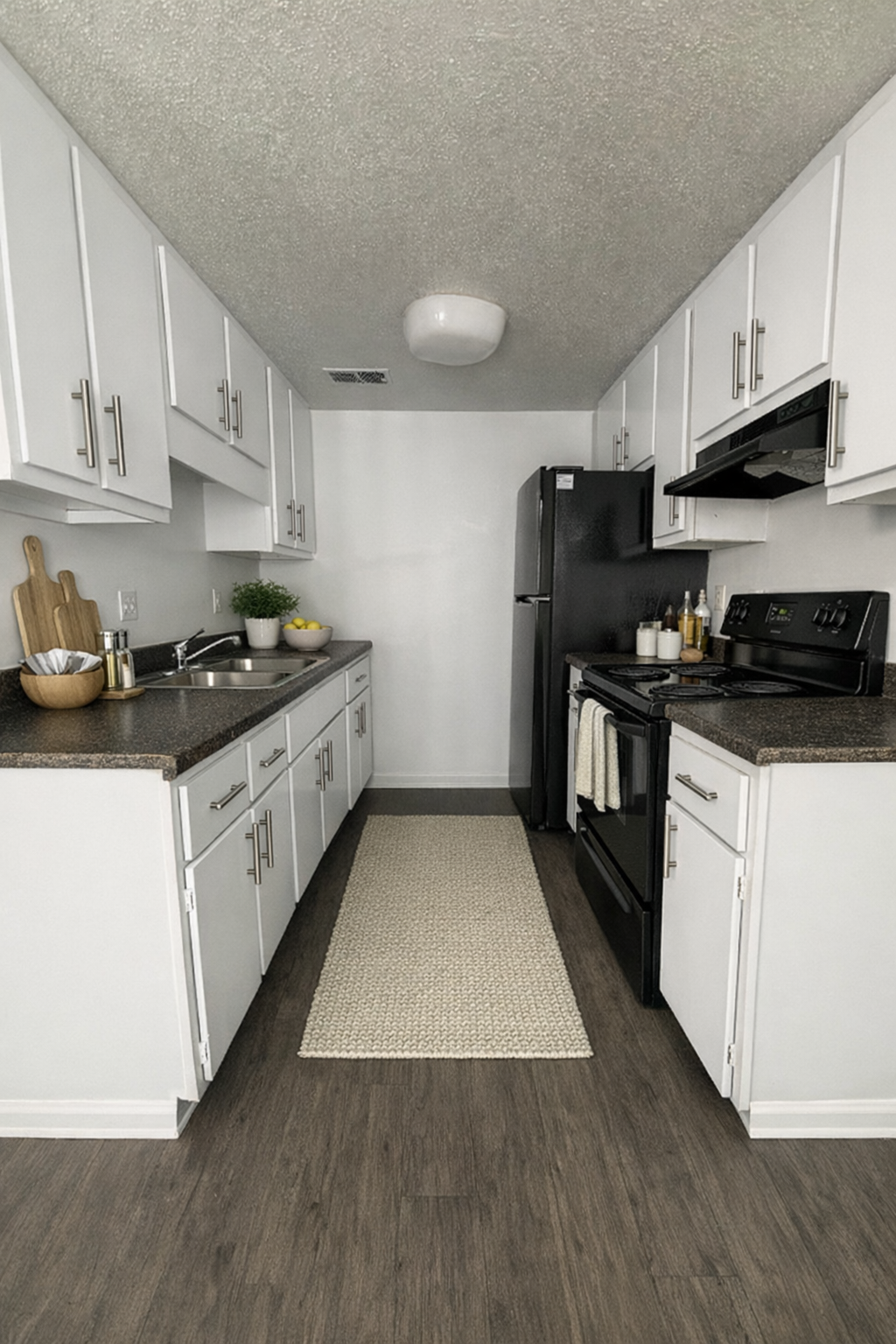 Narrow galley kitchen with white cabinets, black appliances, and a patterned runner on dark flooring.