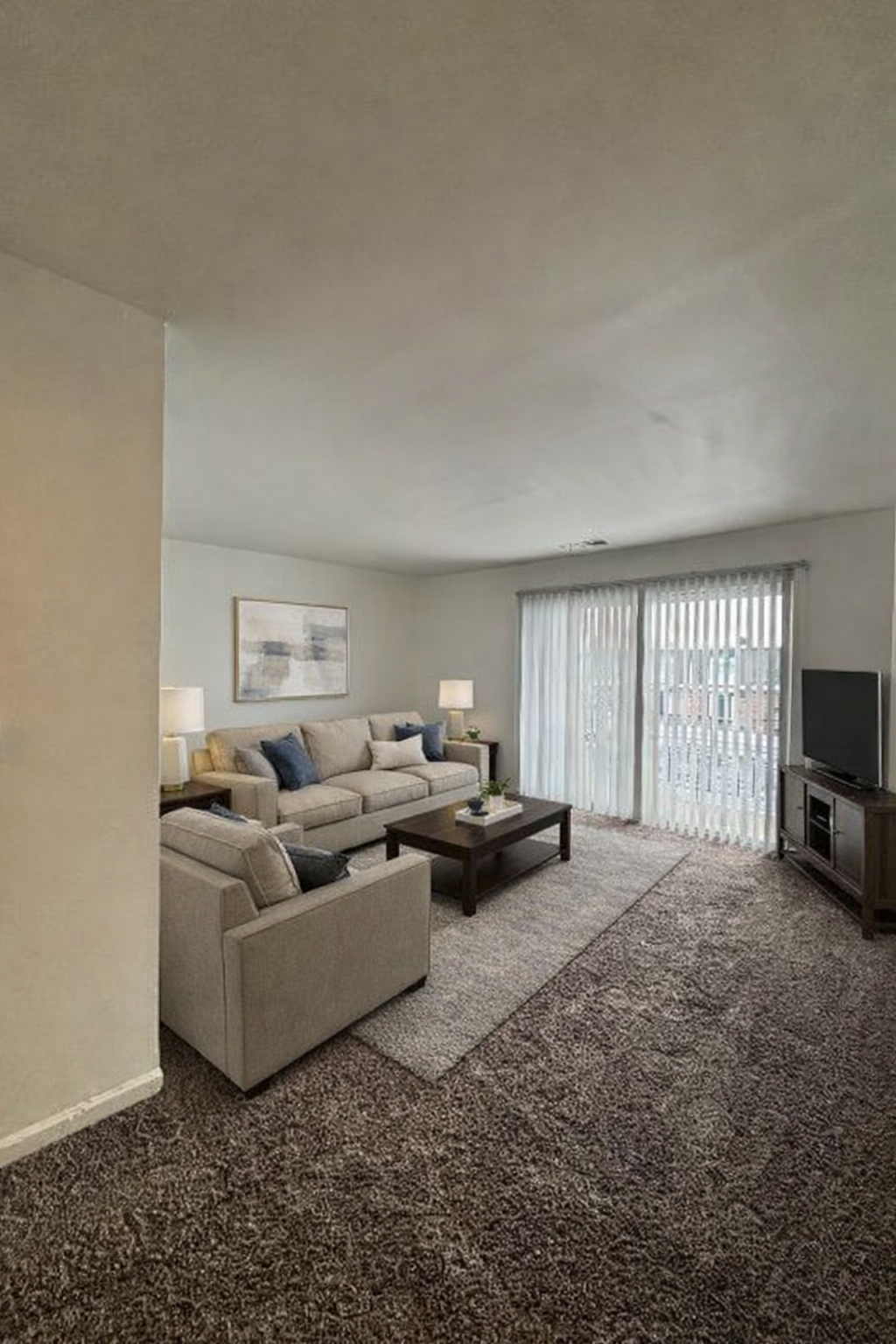 Bright living room with beige sectional sofa, coffee table, TV, and sliding glass doors with vertical blinds.