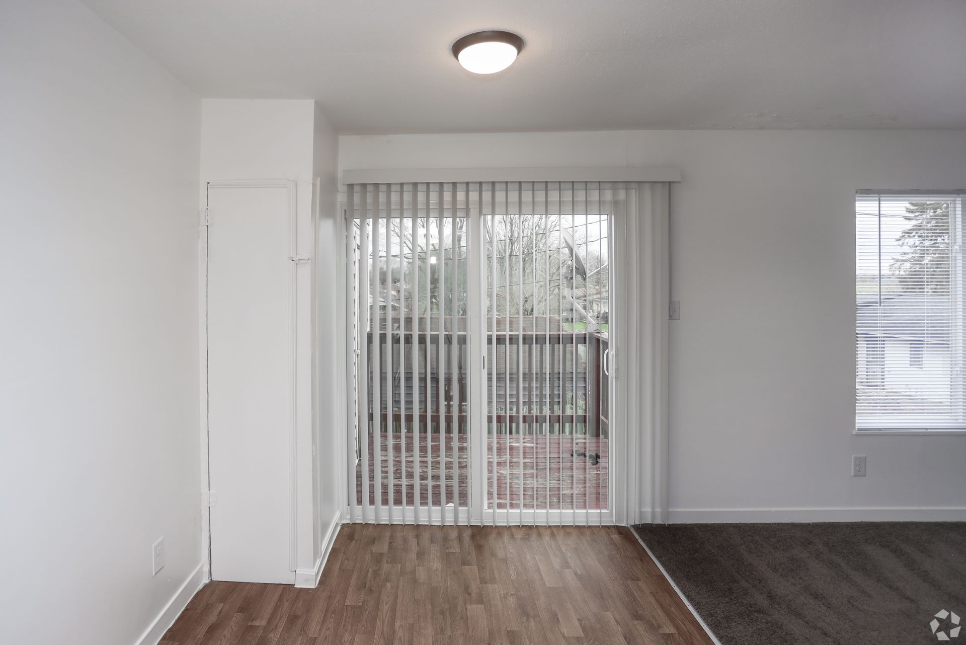 Empty living room with wood floors, white walls, ceiling light, and sliding glass door with vertical blinds