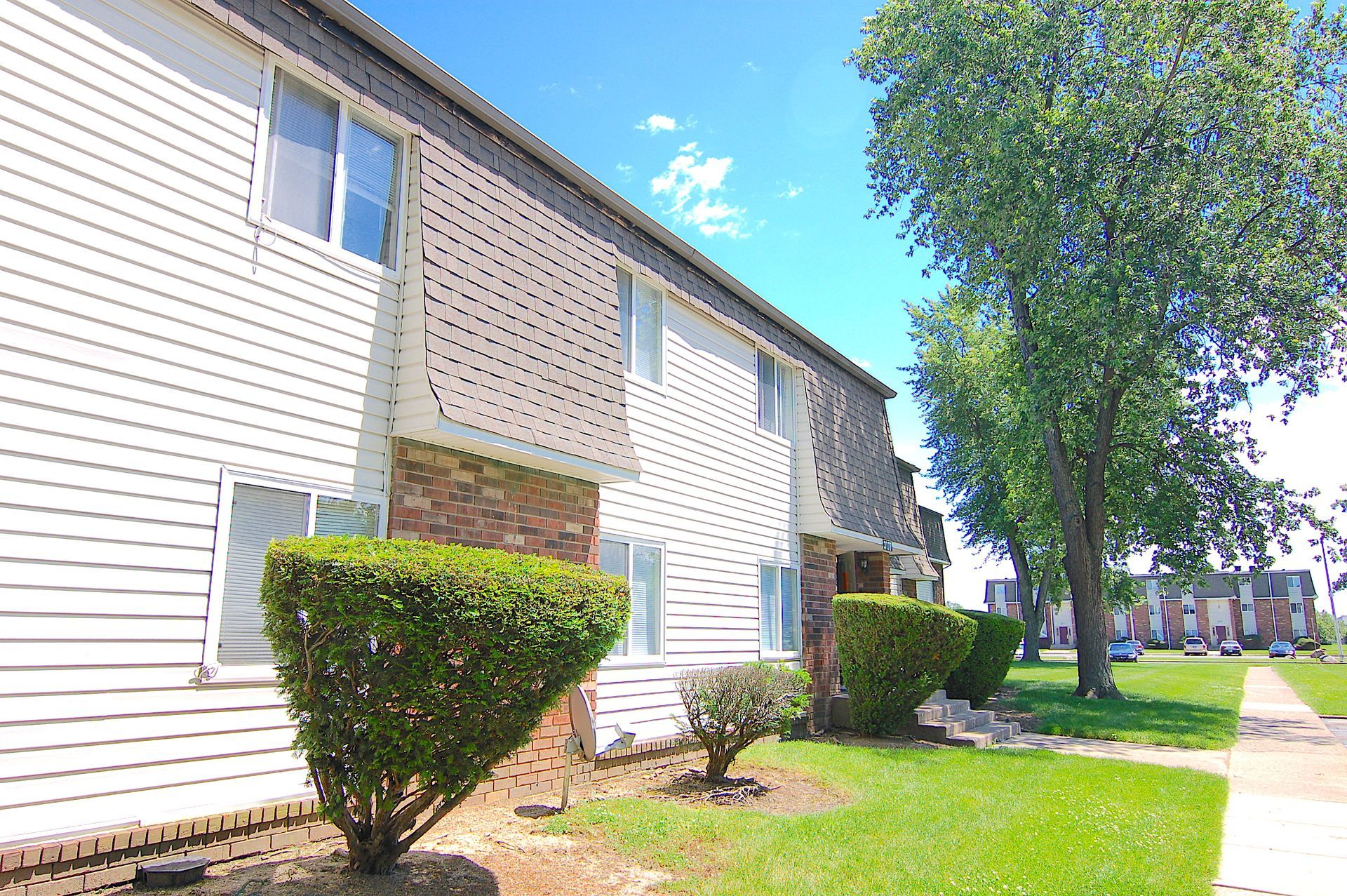 Apartment building exterior with white siding, trimmed hedges, green lawn, and a tree along a sunny sidewalk