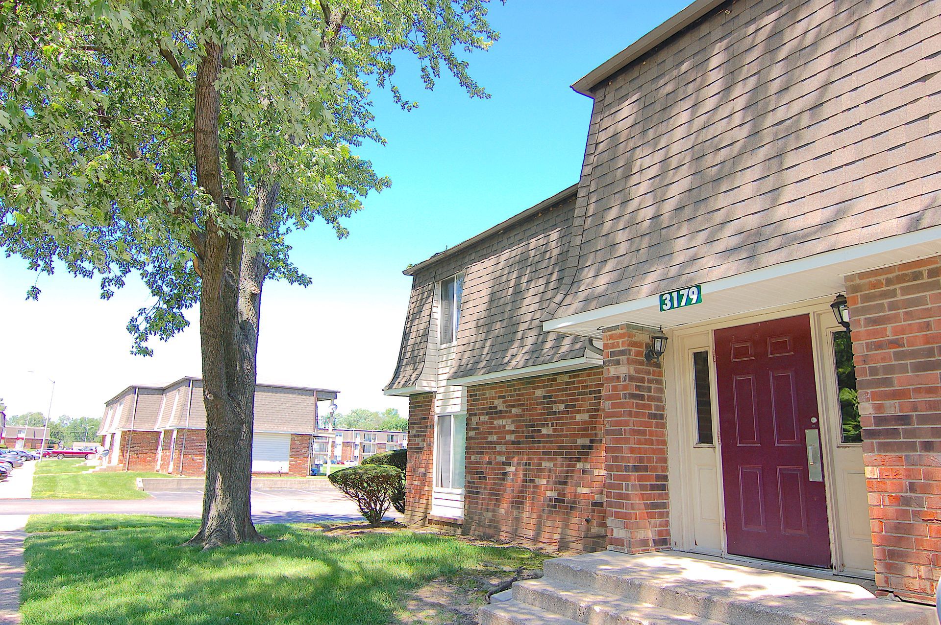 Brick townhouse entrance with red door, tree, and grassy lawn on a sunny day