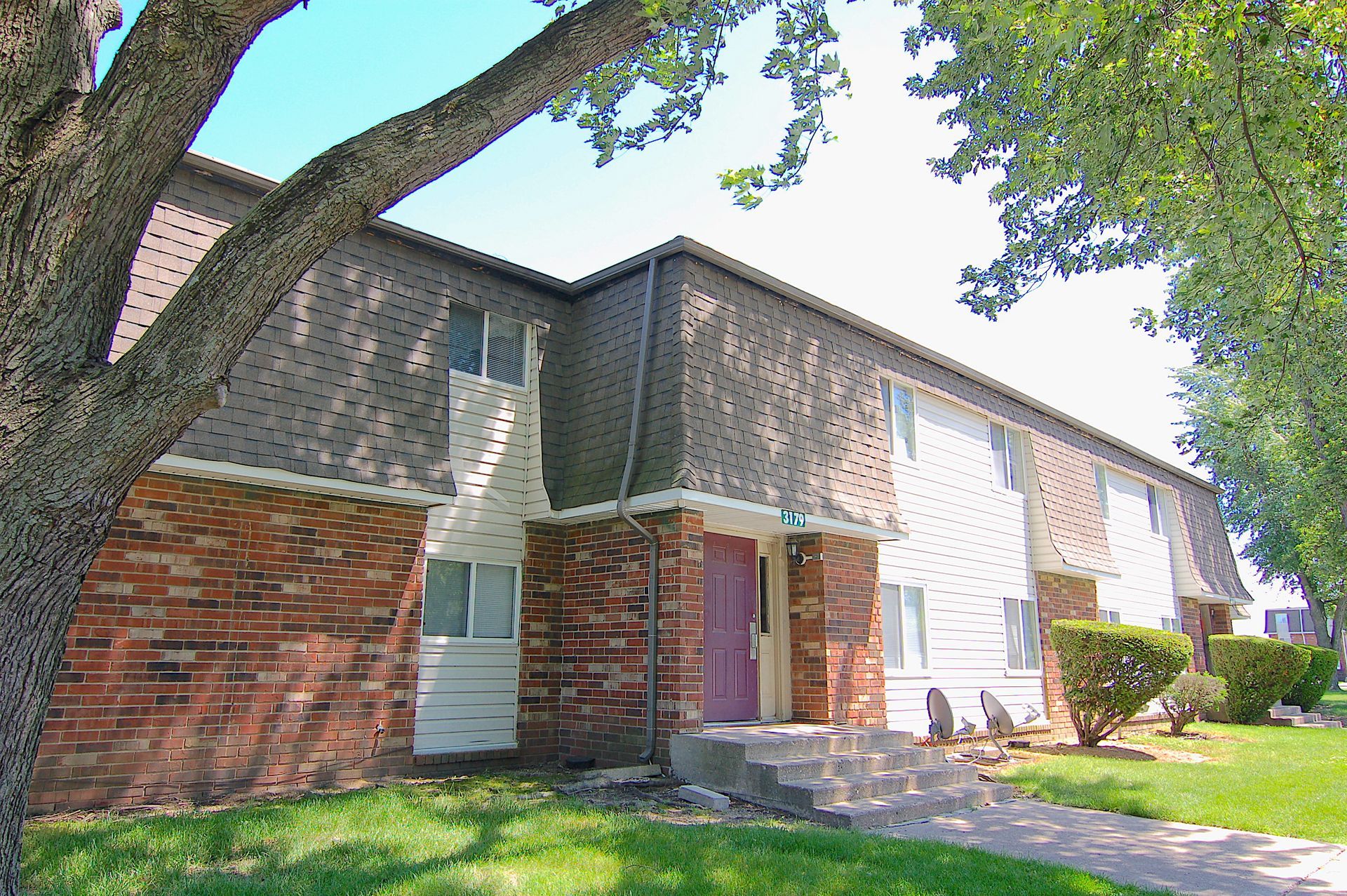 Apartment building exterior with brick and white siding, tree in front, and walkway by the entrance