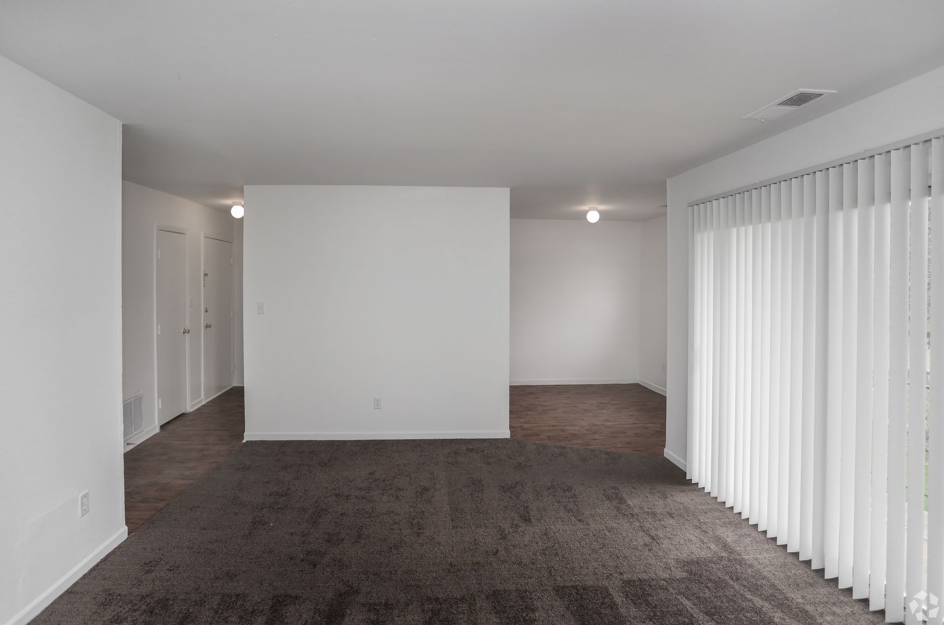 Empty carpeted living room with white walls and vertical blinds along the right side