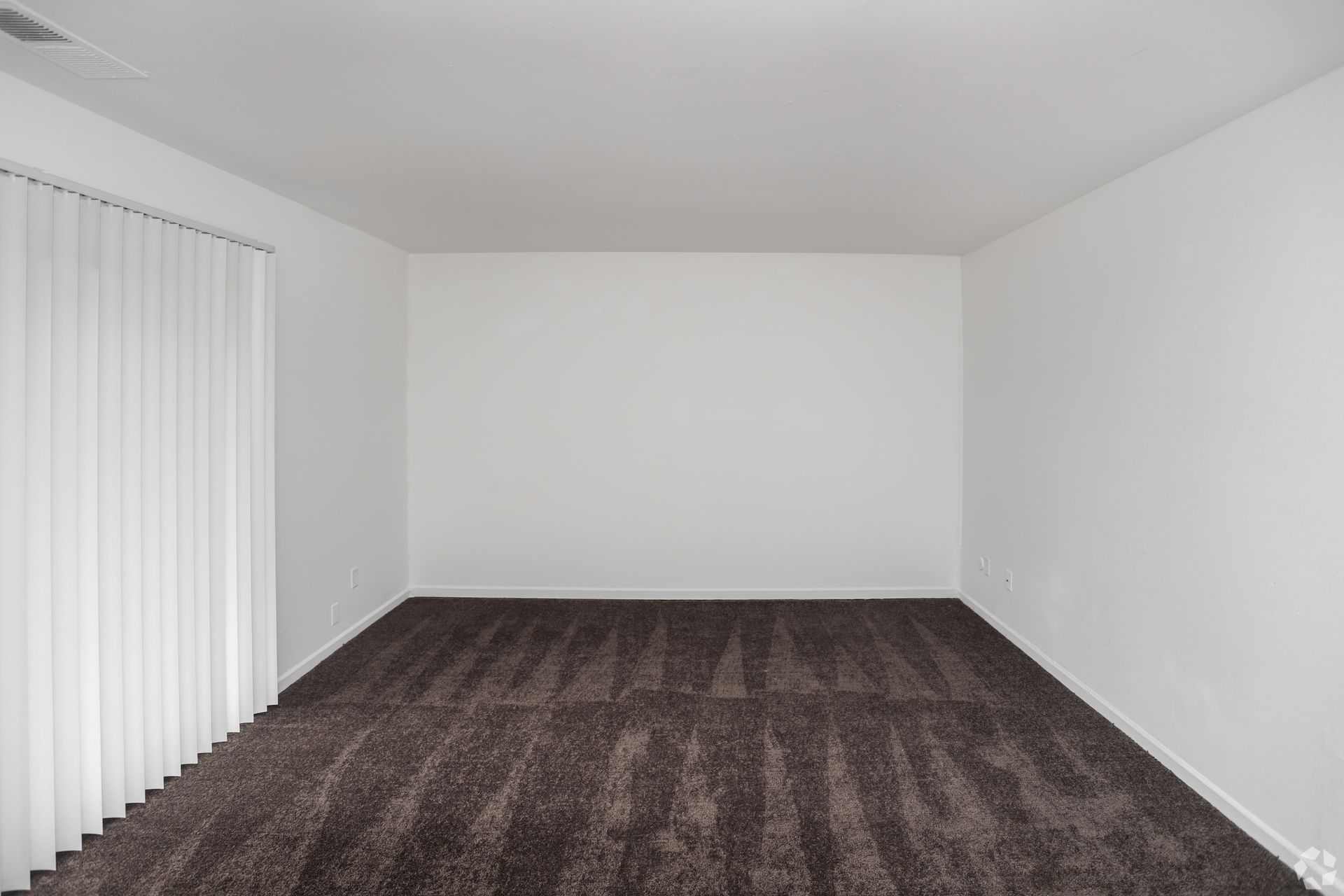 Empty carpeted room with white walls and vertical blinds on the left.