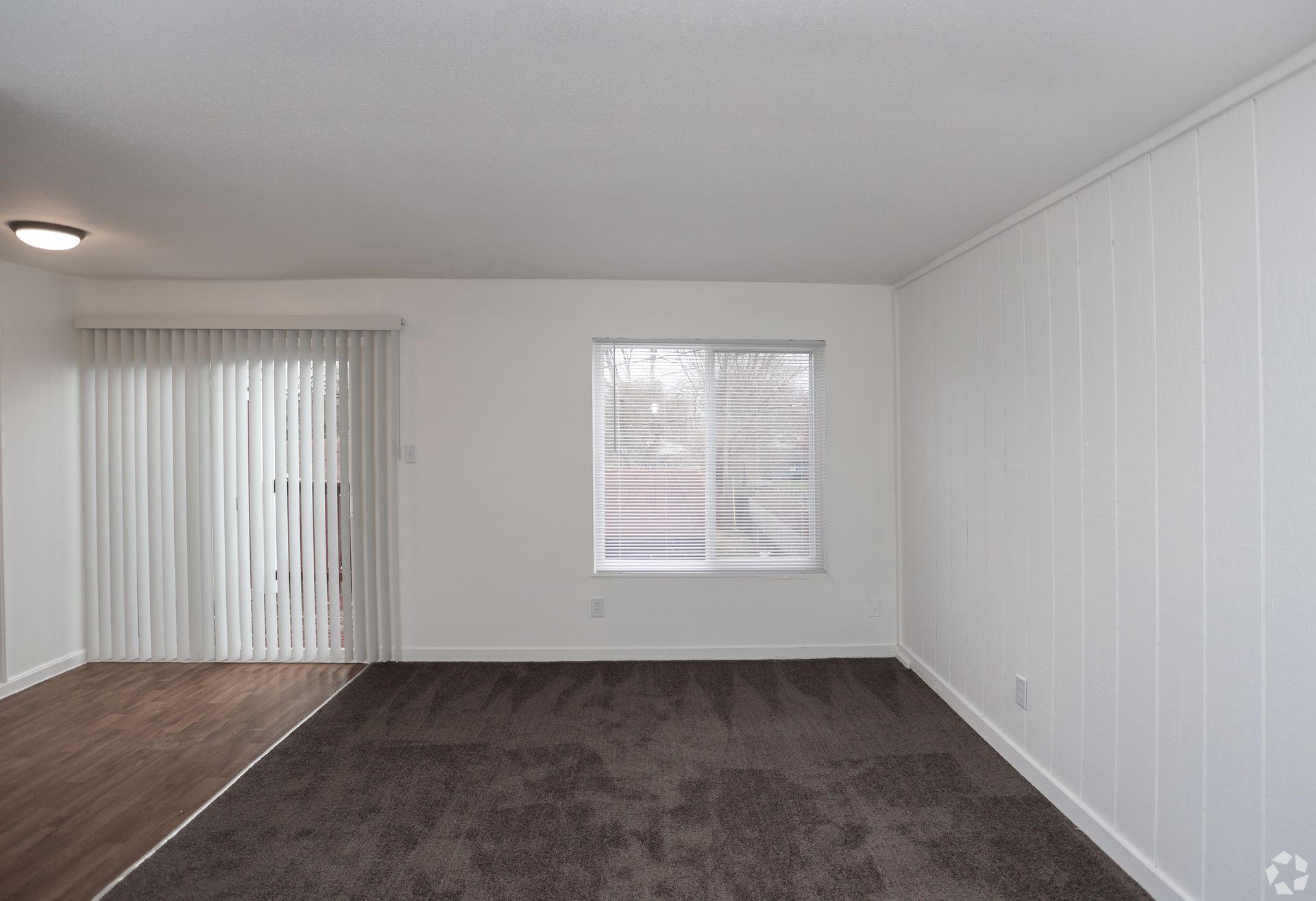 Empty living room with gray carpet, white walls, sliding glass door, and window with blinds.