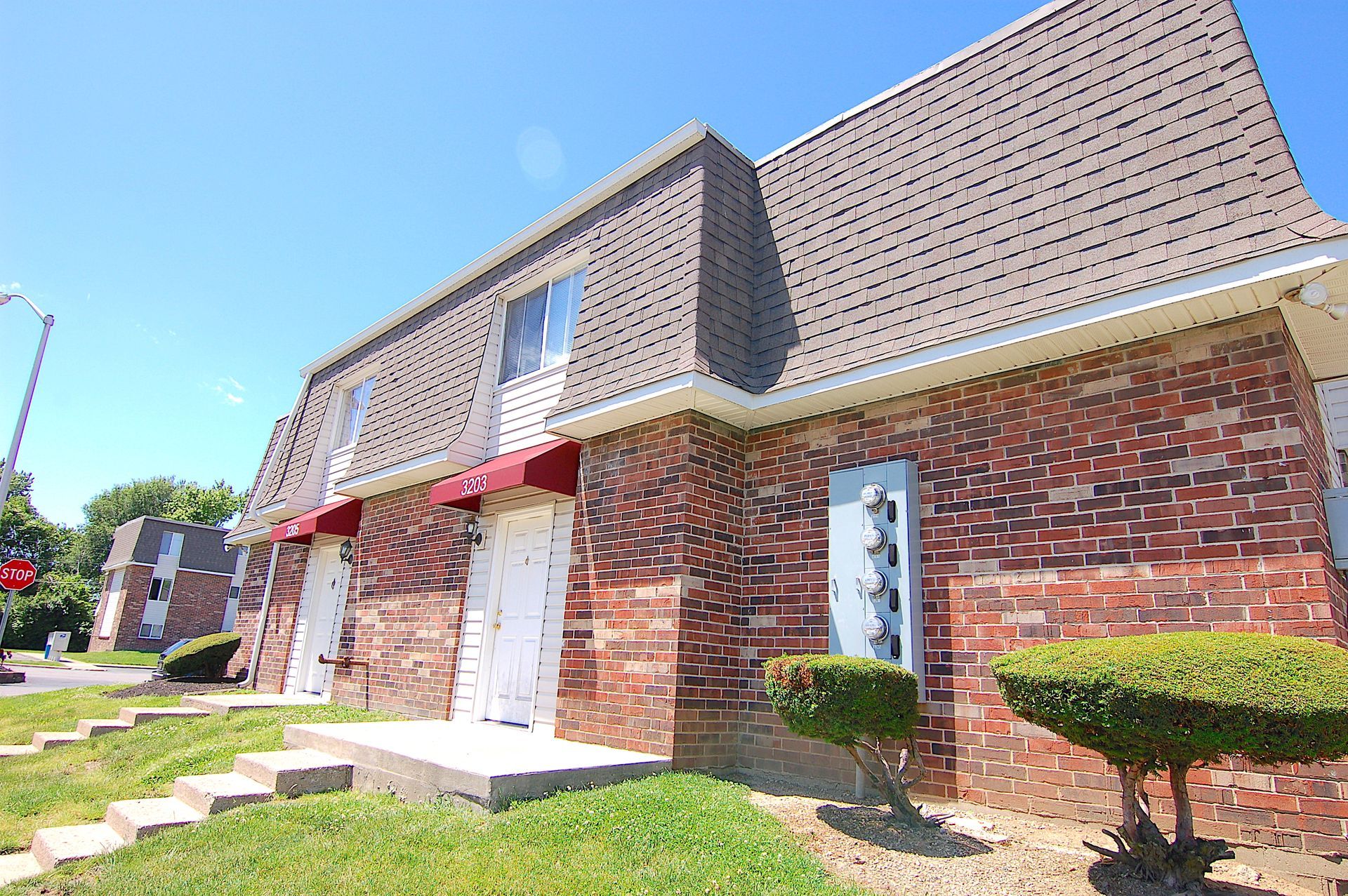 Brick apartment building with red awnings, white doors, and neatly trimmed shrubs under a bright blue sky
