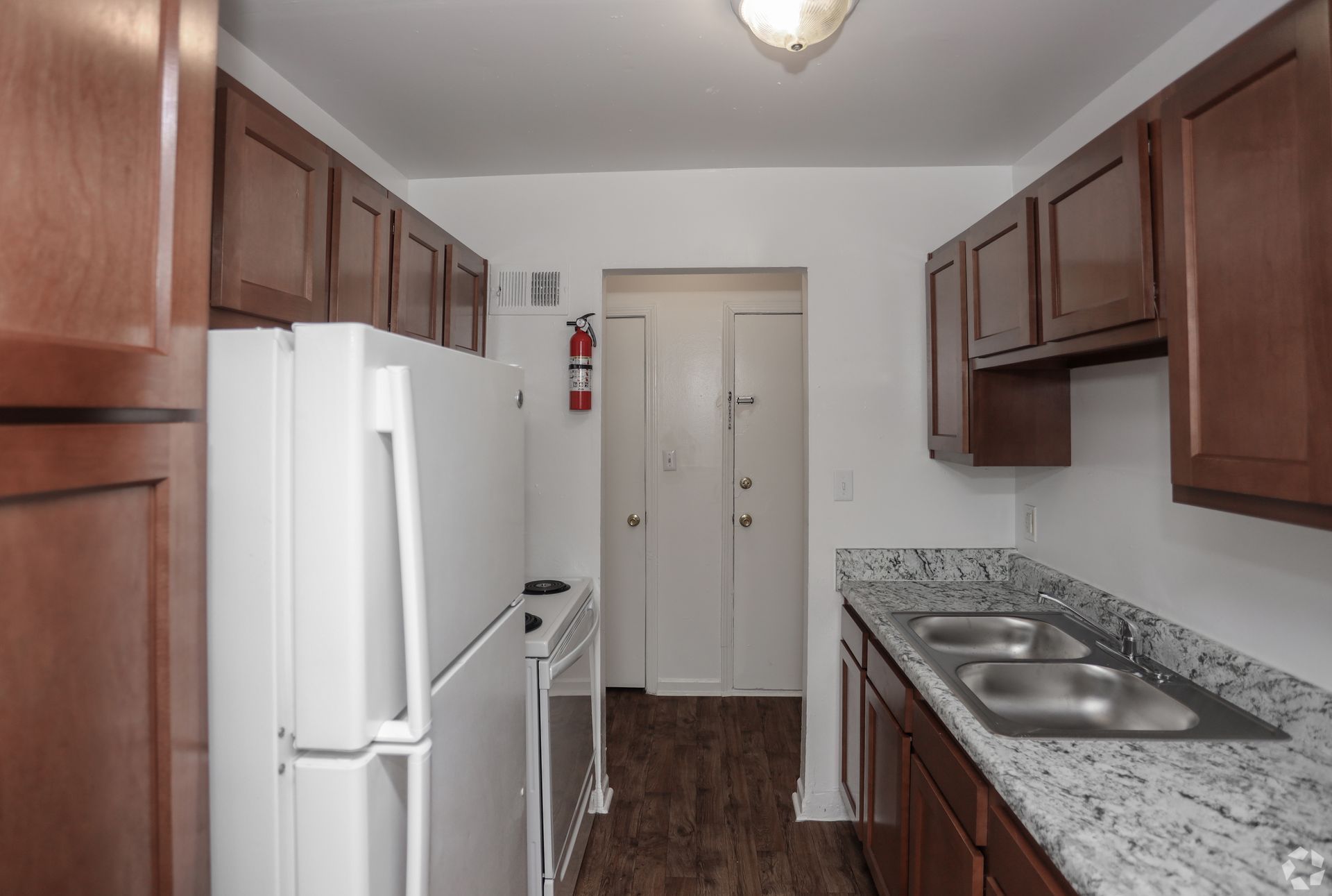 Narrow kitchen with white fridge, brown cabinets, granite counters, and a doorway at the end.