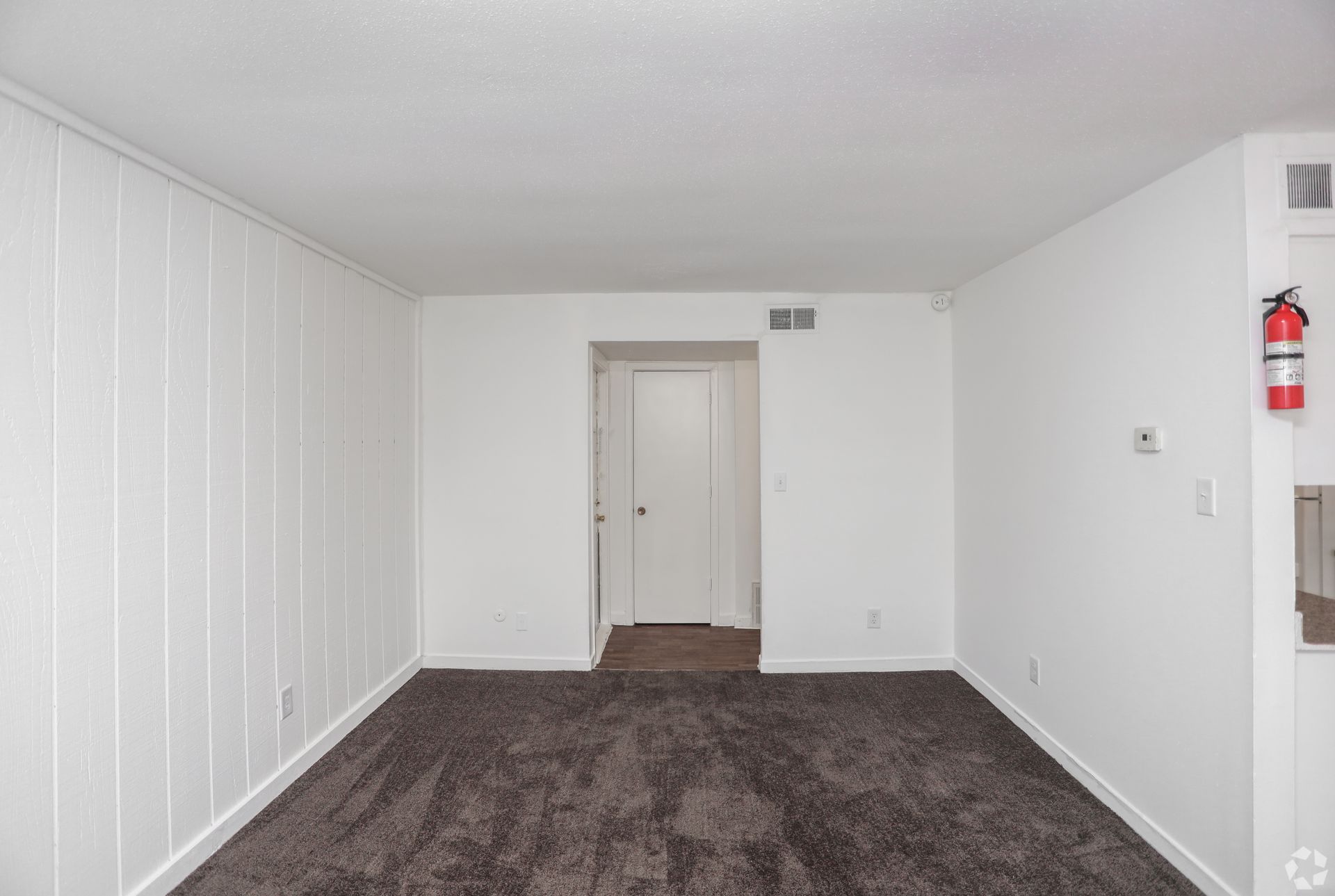 Empty white room with dark carpet, paneled wall, open doorway, and fire extinguisher on the right wall