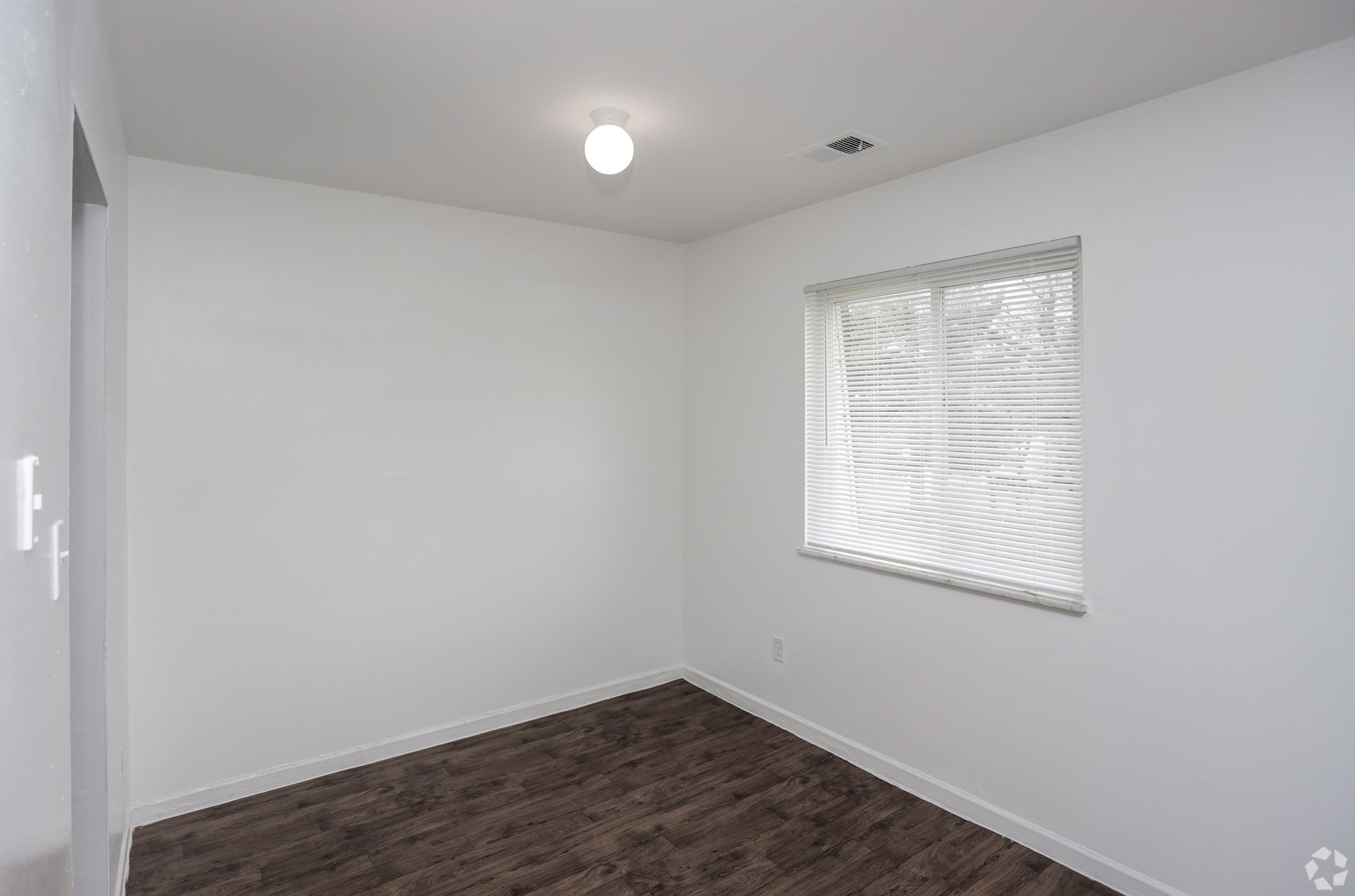 Empty white room with dark wood flooring, a ceiling light, and a small window with blinds.