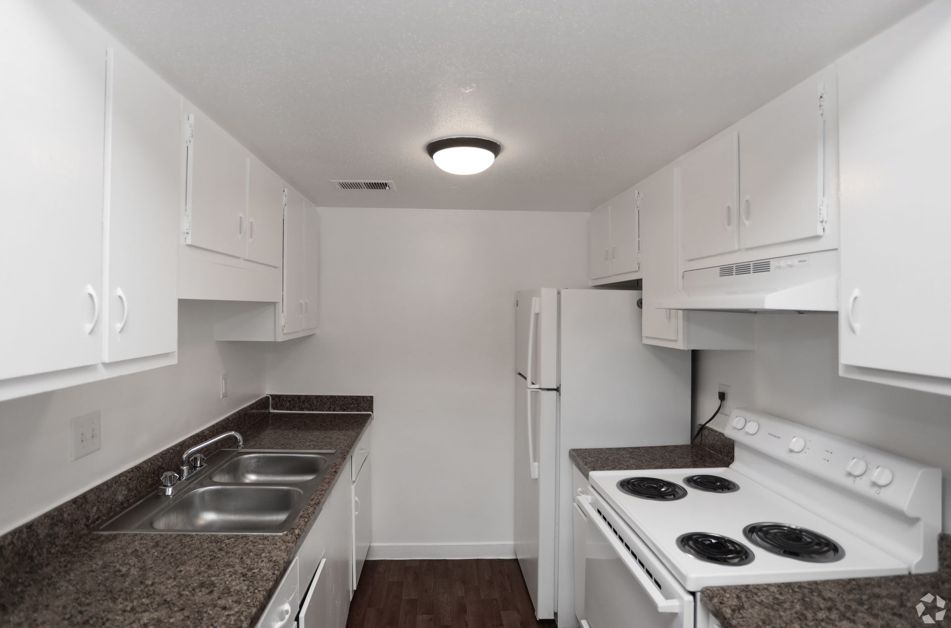 Bright galley kitchen with white cabinets, dark counters, stove, sink, and refrigerator.