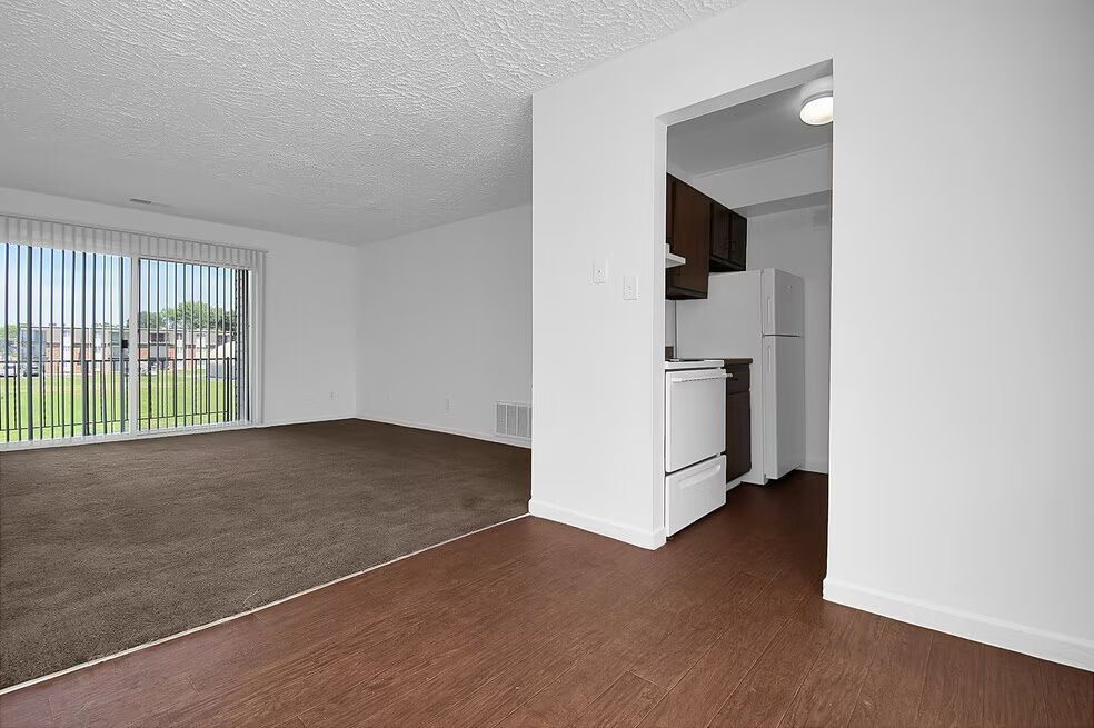 Empty apartment living room with brown carpet, white walls, and a kitchen alcove visible.