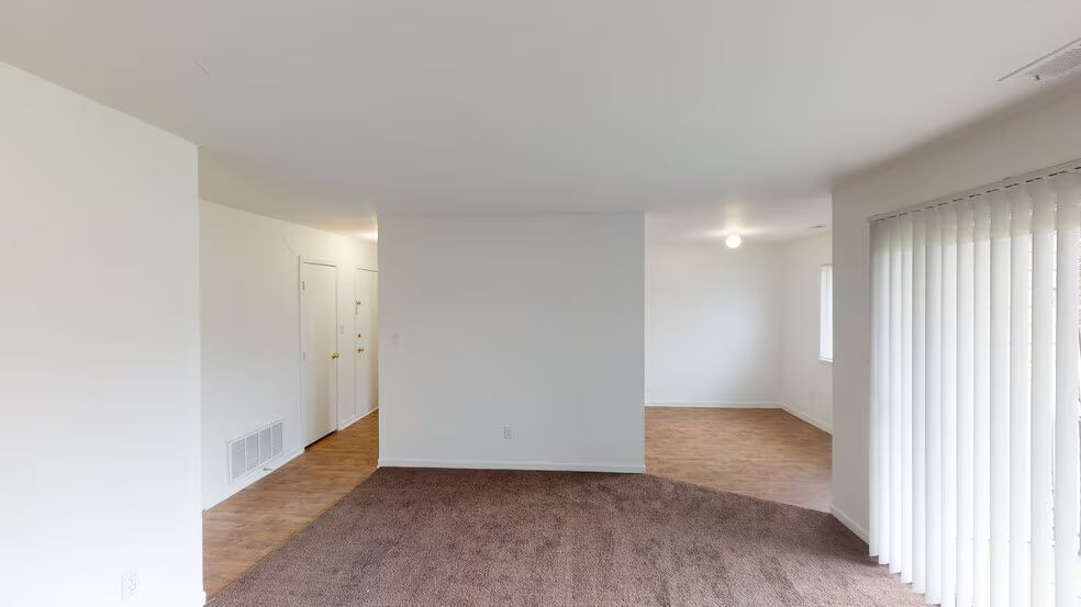 Empty carpeted living room with white walls, vertical blinds, and open doorway to a hallway