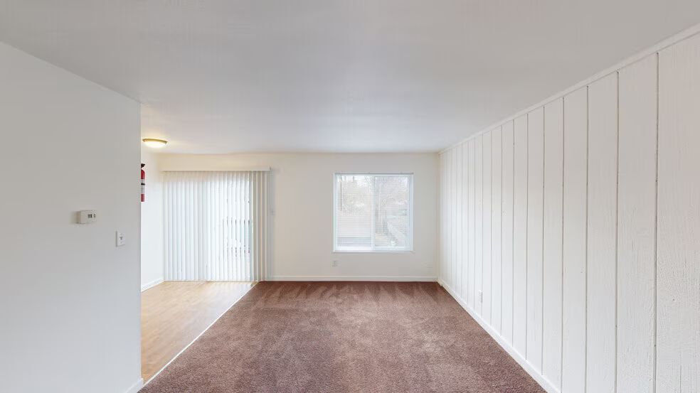 Empty living room with brown carpet, white walls, vertical blinds, and a window letting in light