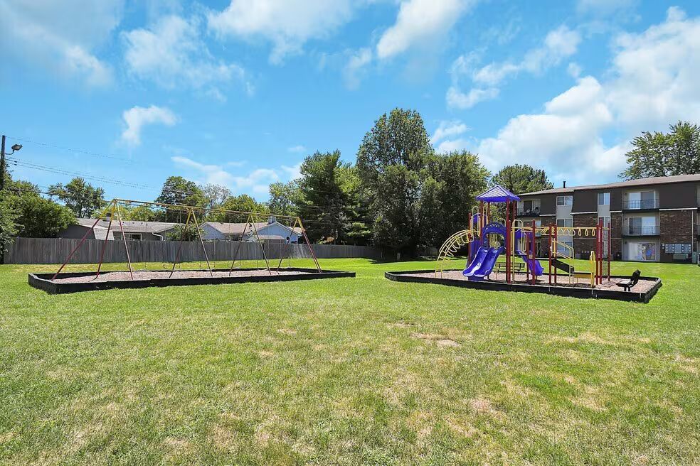 Playground with purple equipment in a grassy park under a blue sky, beside apartment buildings