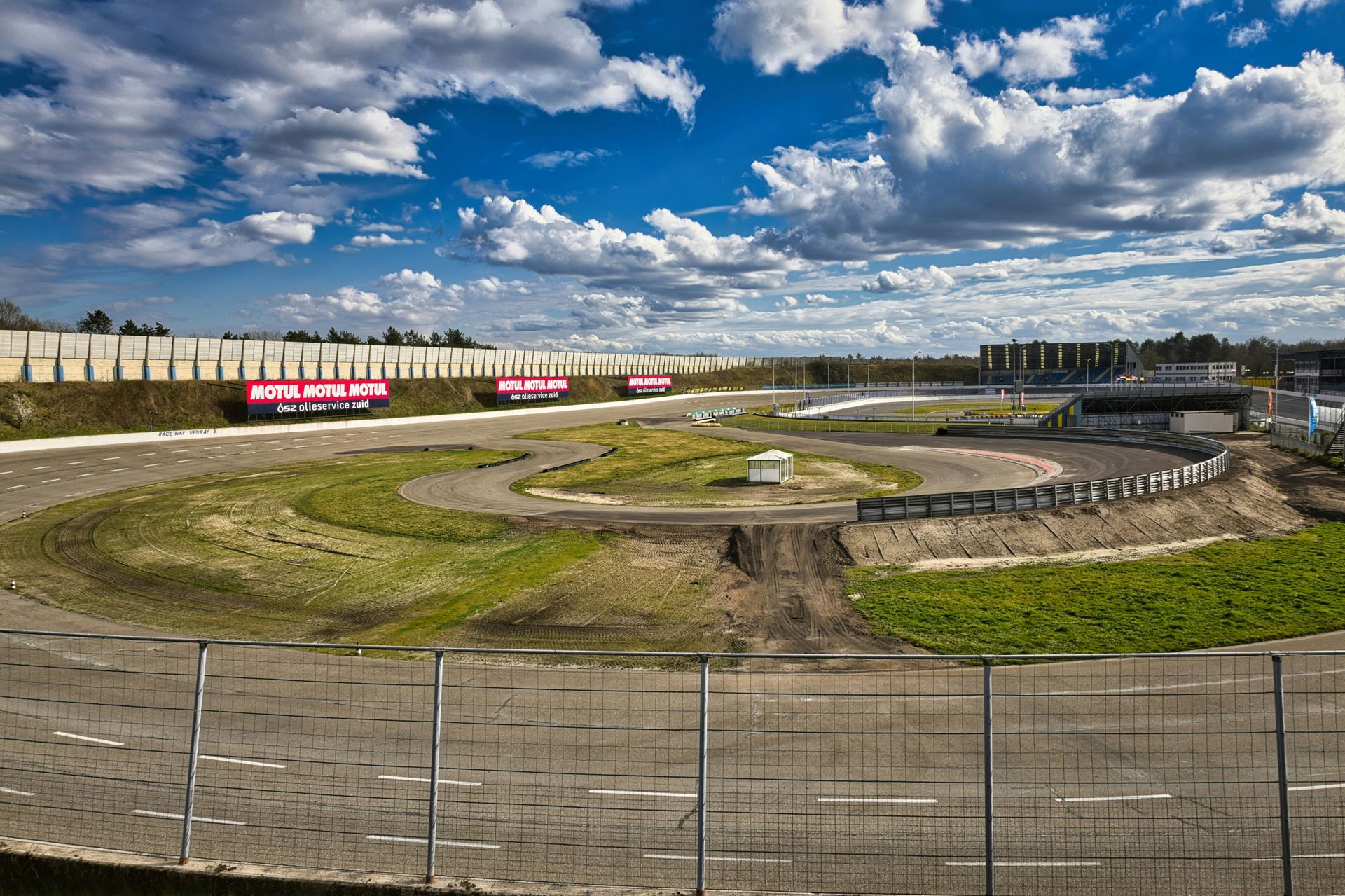 Wide dirt racetrack under a cloudy blue sky, seen through a fence with banners along the far side