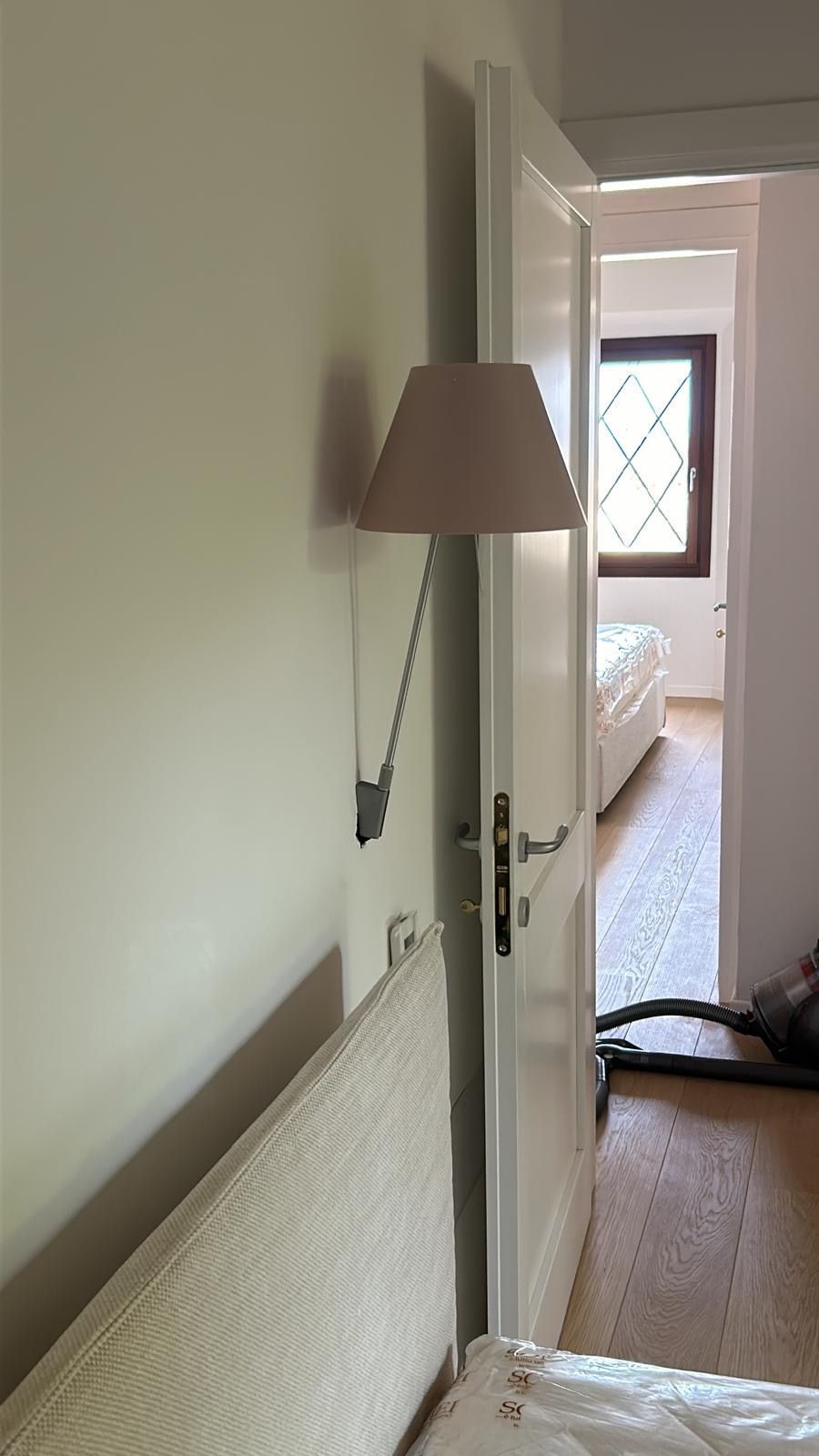 Stretto corridoio della camera da letto con porta aperta, lampada da parete e letto visibile sullo sfondo.