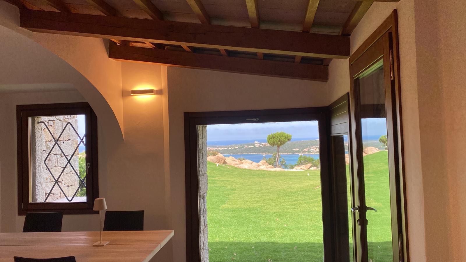 Accogliente camera con porta e finestra aperte che incorniciano un prato verde brillante e la vista sul lago in lontananza.