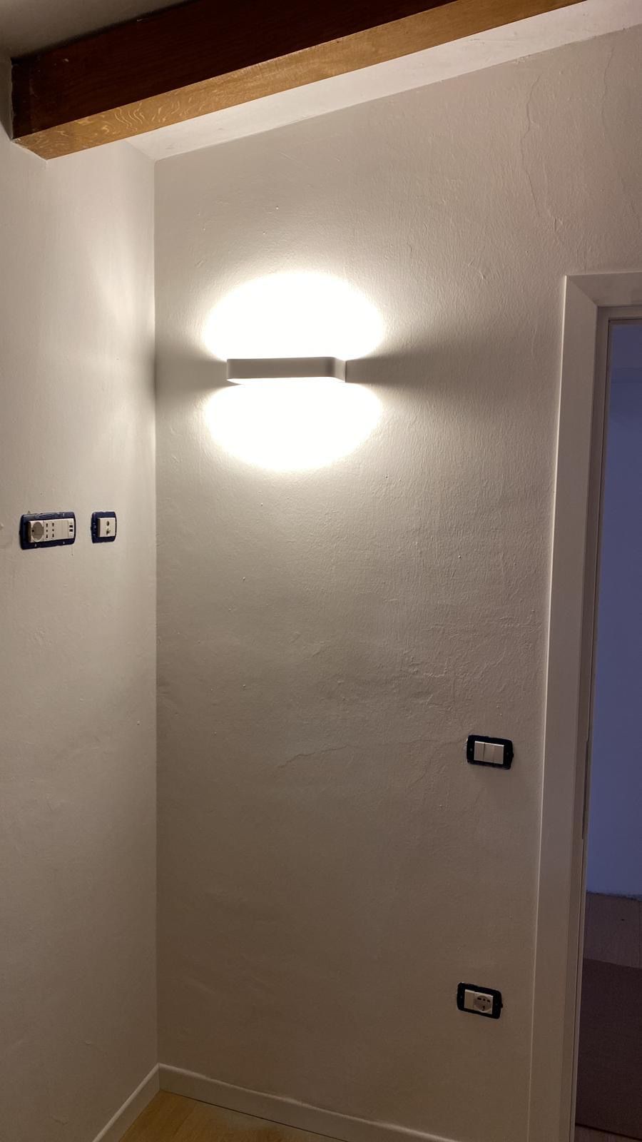 Parete del corridoio con applique luminosa, due piccoli apparecchi a parete e una porta sulla destra.