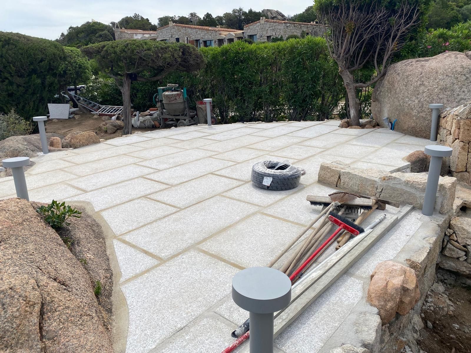 Patio in pietra in costruzione con grandi lastre bianche, attrezzi e pali di bordatura in un contesto di giardino.