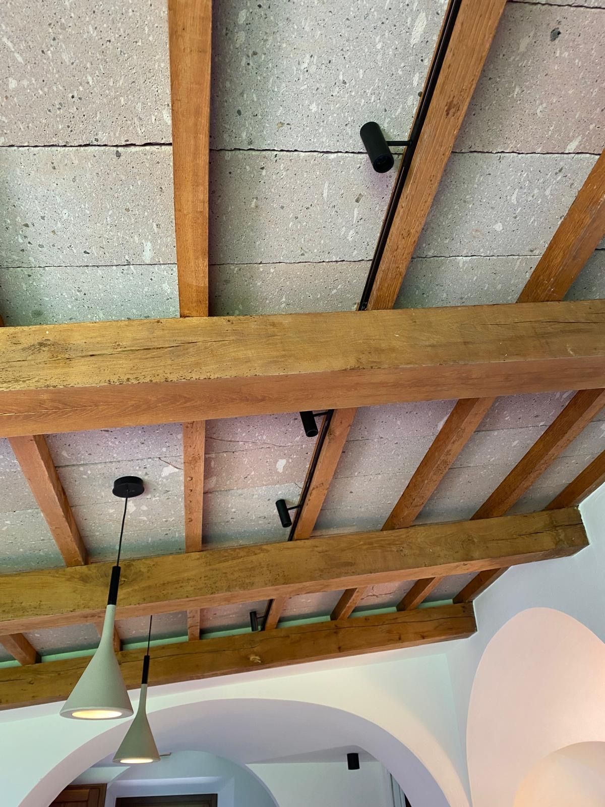 Travi a vista in legno con pannelli acustici grigi, lampade a sospensione e pareti ad arco bianche sottostanti.