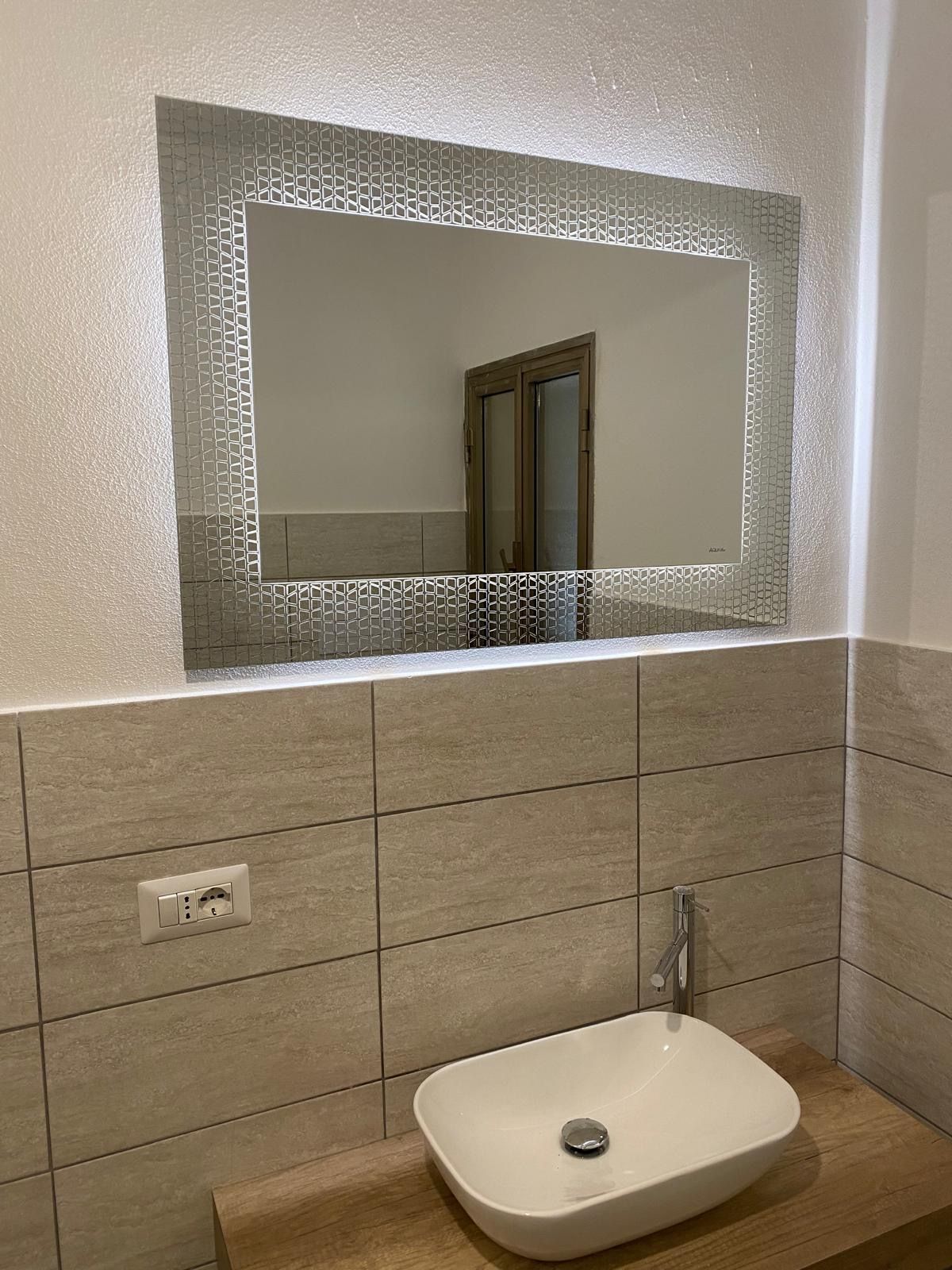 Moderno lavabo da bagno con bacinella bianca e specchio rettangolare retroilluminato sopra una parete piastrellata beige.