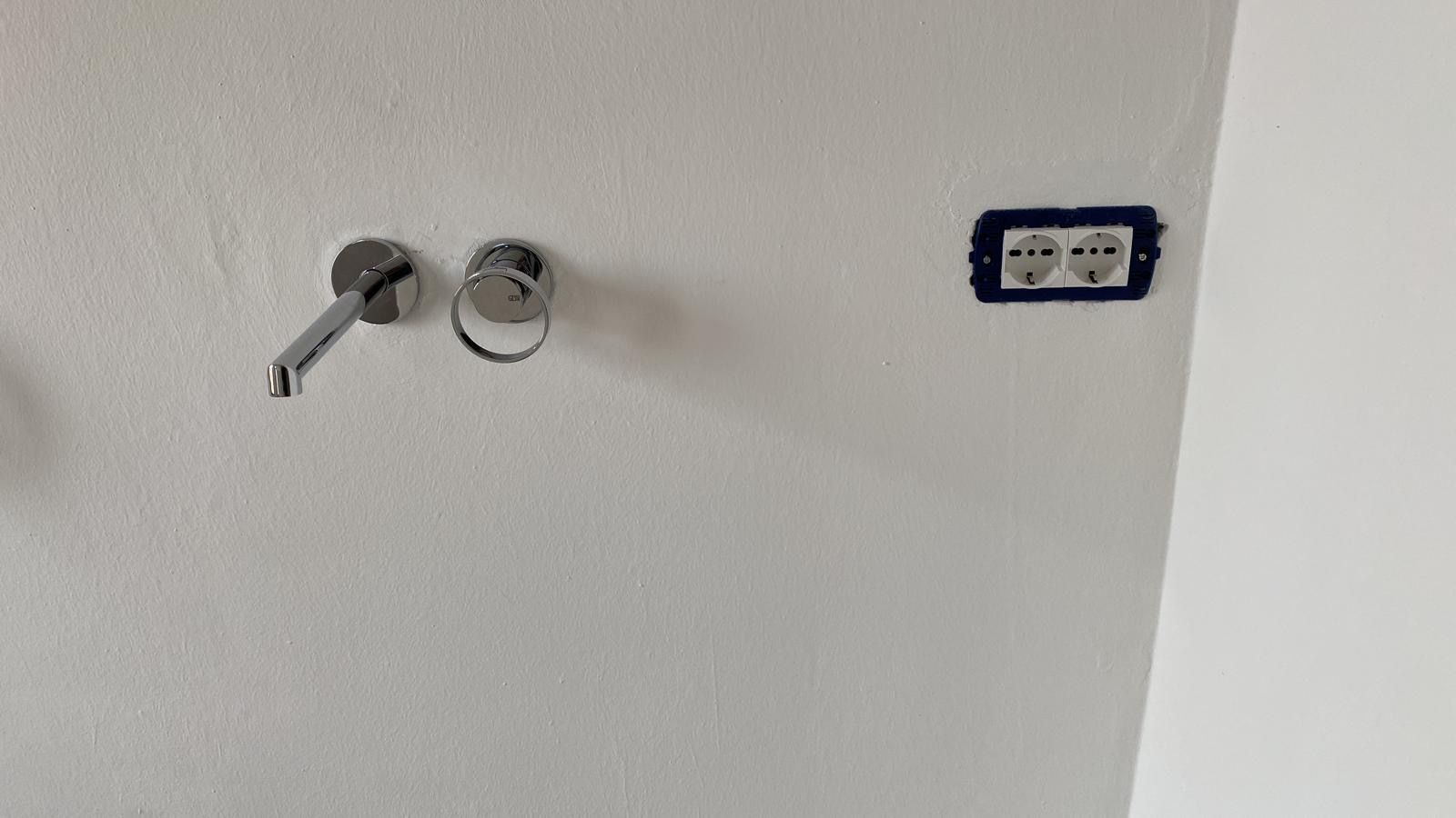 Parete bianca con due rubinetti a vista e una placca di presa elettrica blu nelle vicinanze.
