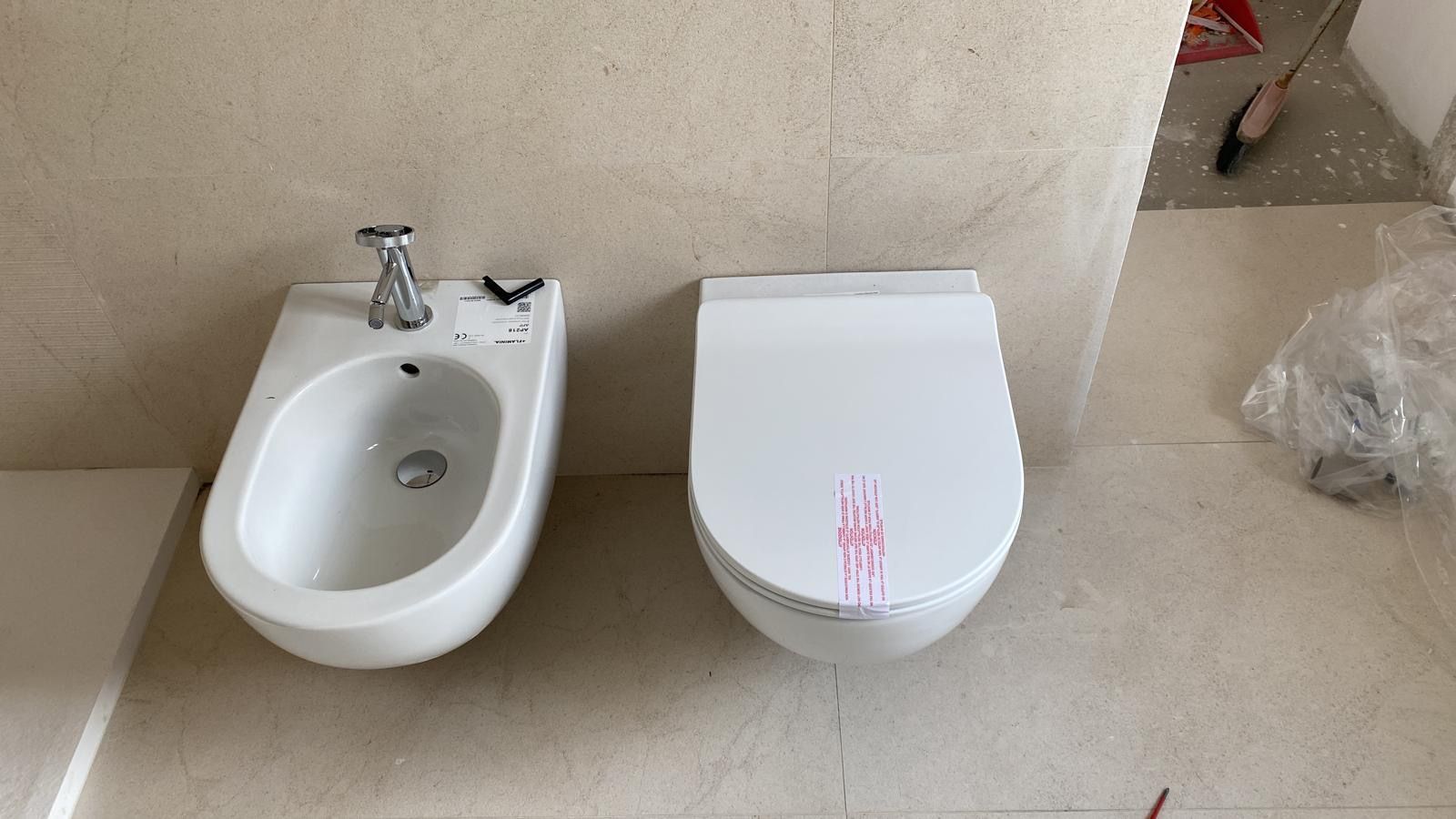 Un piccolo bidet e un WC bianchi affiancati su un pavimento del bagno piastrellato di beige.