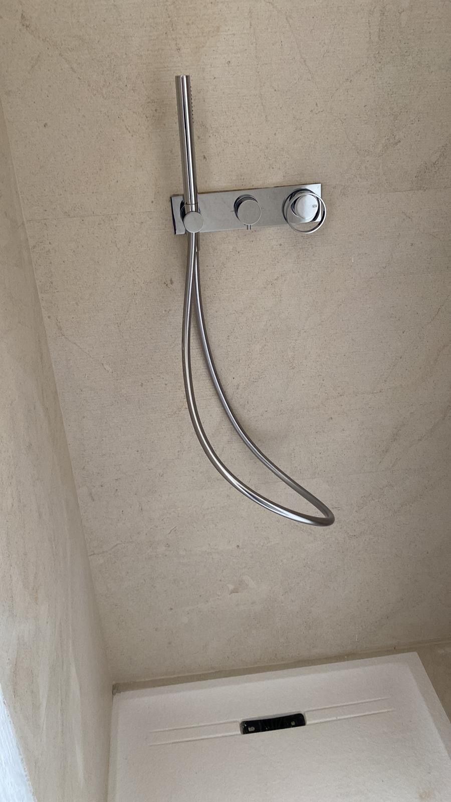 Maniglione di sicurezza cromato per doccia con impugnatura curva, montato su una parete beige sopra una panca da doccia.